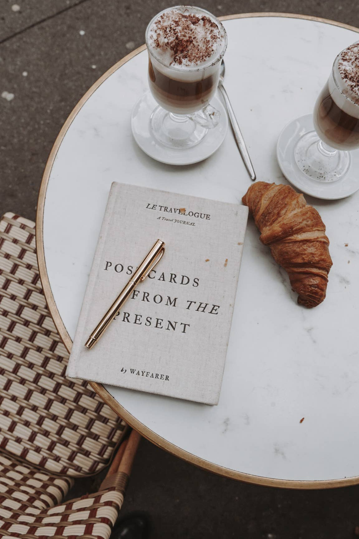 Book titled 'Postcards from the Present' with a pen and croissant on a marble table.