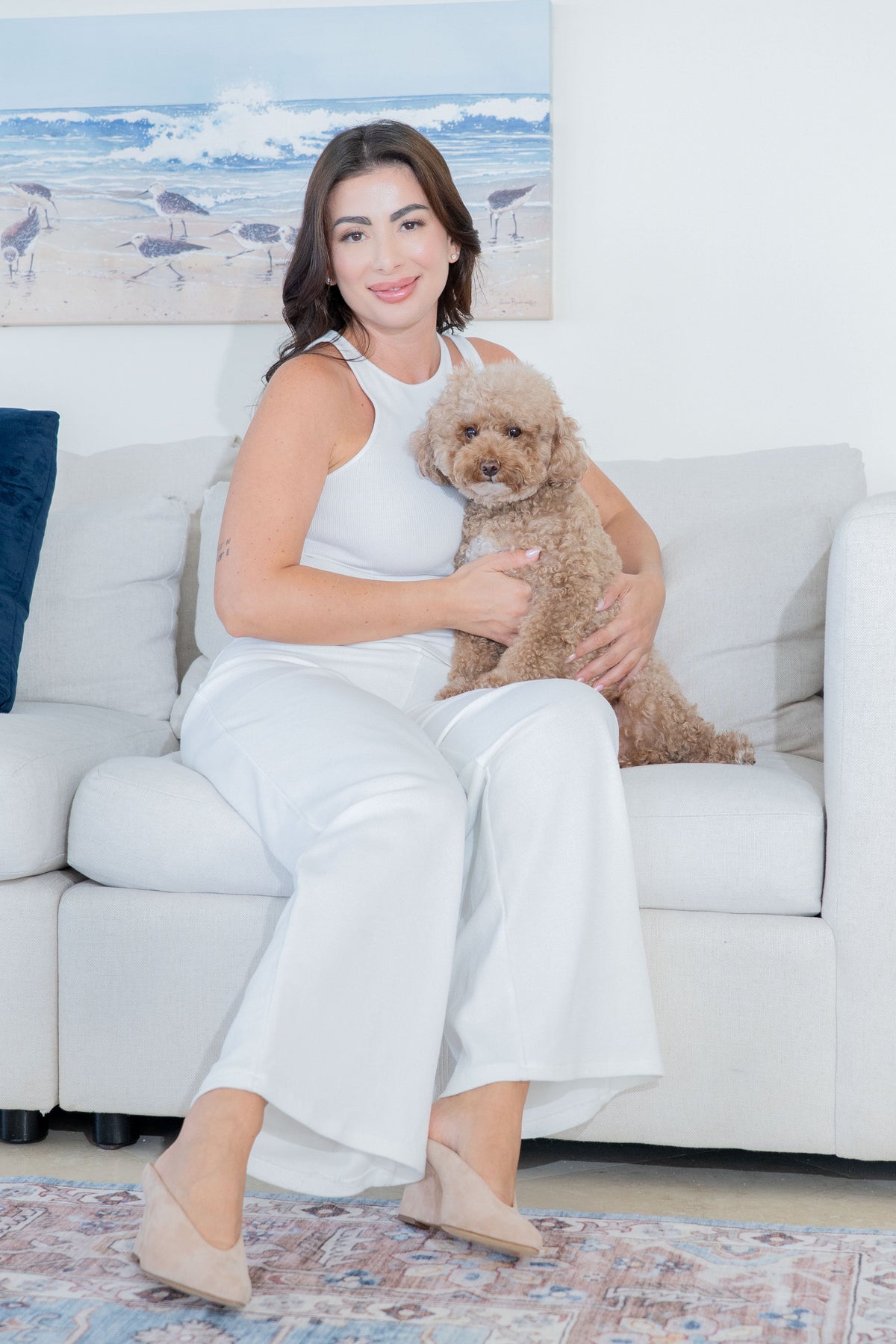 A woman in a white sleeveless top, white wide-leg pants, and suede beige wedged heels sits on a white sofa holding a small fluffy tan dog. The room has a light wall with a beach painting above the sofa, and a patterned rug is visible on the floor.