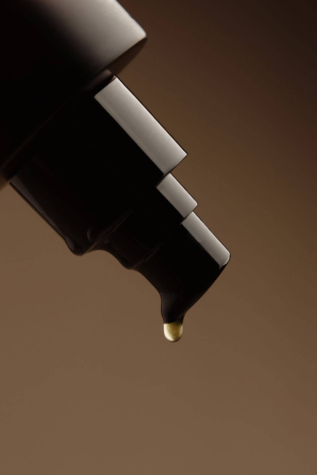 A close-up of a black cosmetic dropper bottle tip with a single gold liquid droplet hanging over a smooth brown background.