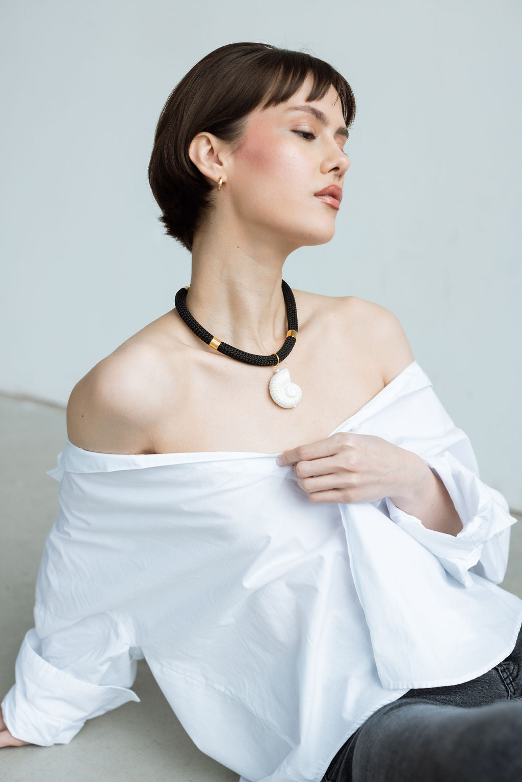 Woman wearing a white off-shoulder top and a black necklace with a large pendant against a light gray background.