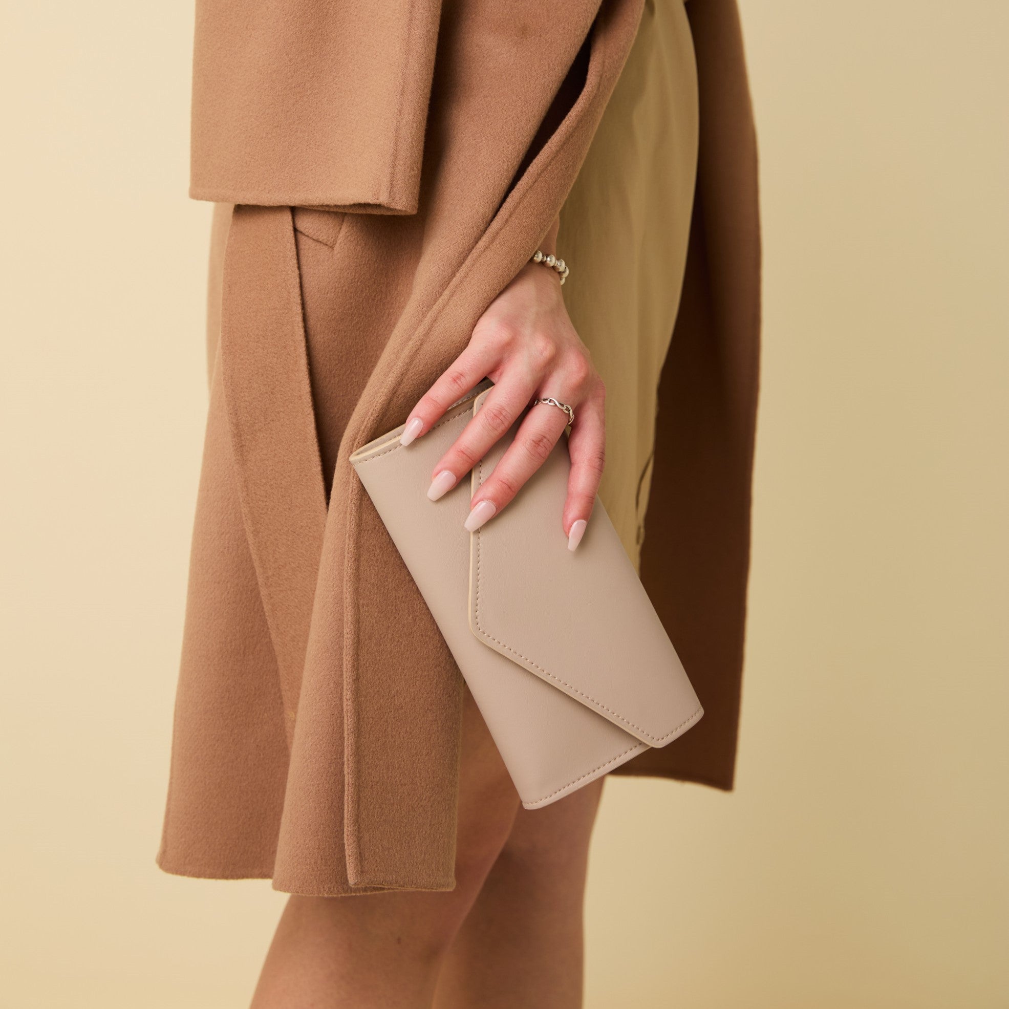 Person wearing a brown coat holding a beige clutch against a beige background