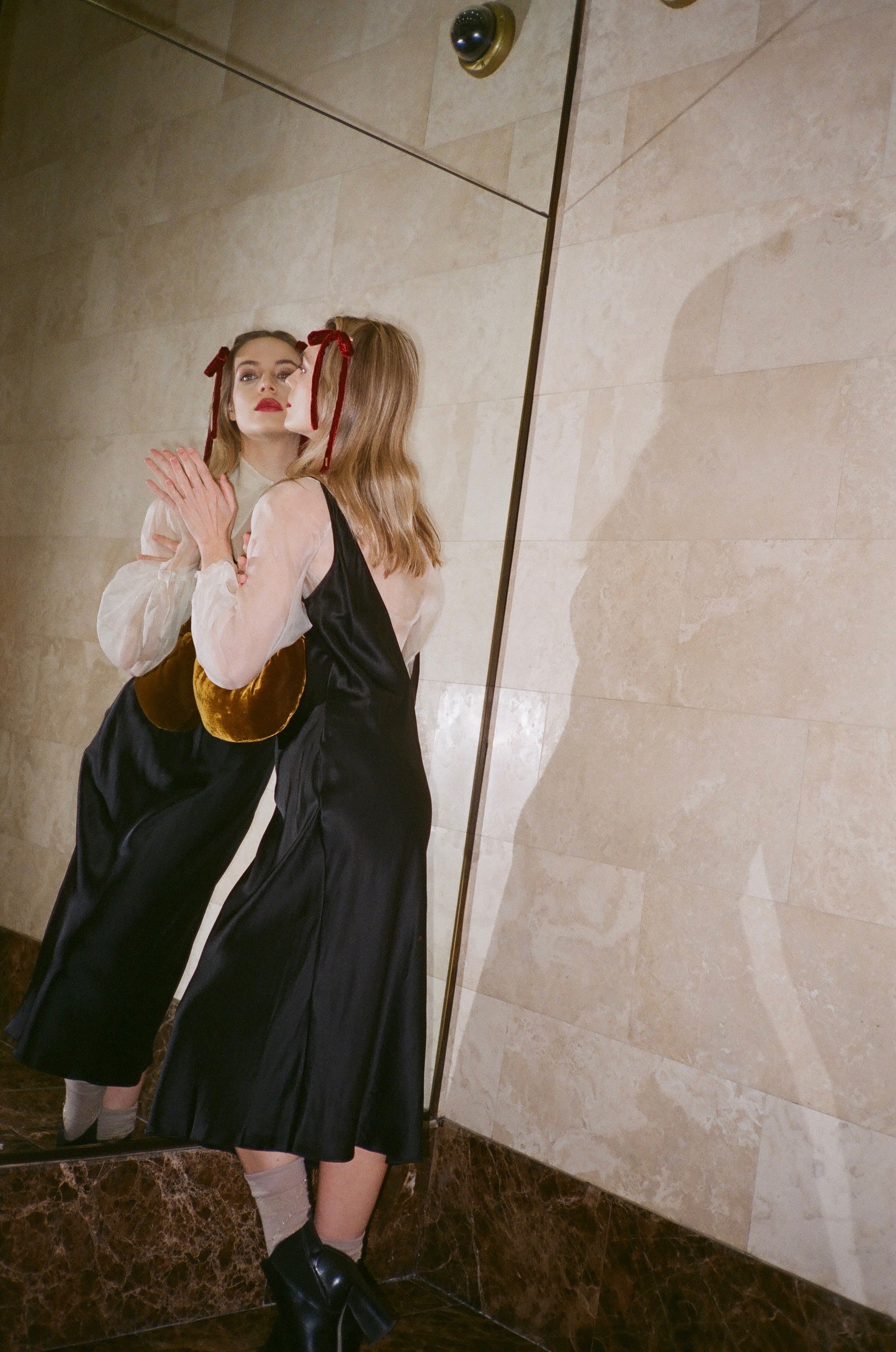 A woman wearing a black silk slip dress over a sheer white silk blouse with red ribbon velvet headbands, standing in front of a large mirror in a tiled room.