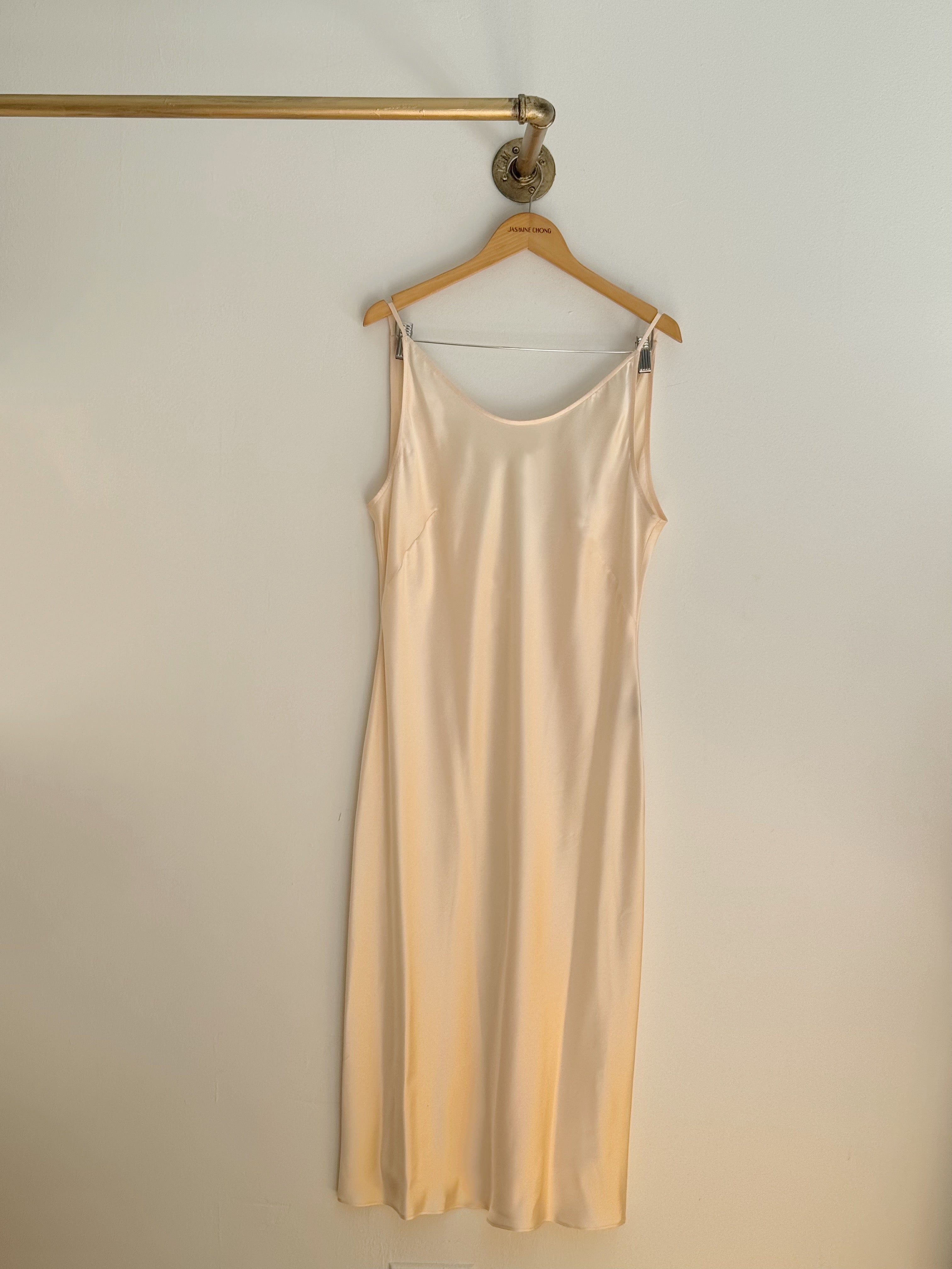 A light beige satin silk slip dress hanging on a wooden hanger from a gold wall rod against a plain off-white wall.