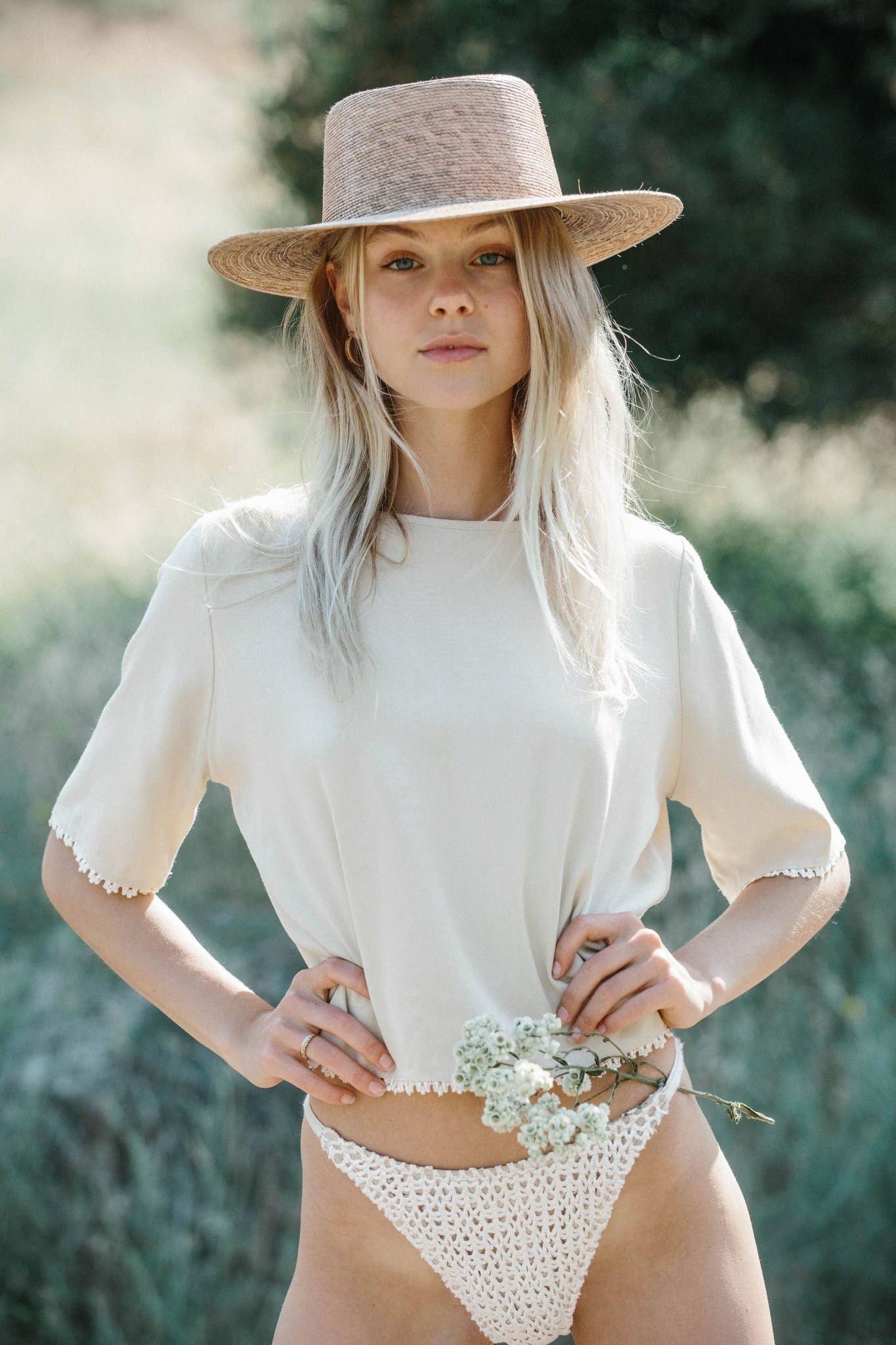 A woman wearing a beige Magnolia Blouse with lace trim and a straw hat, standing in a natural outdoor setting.