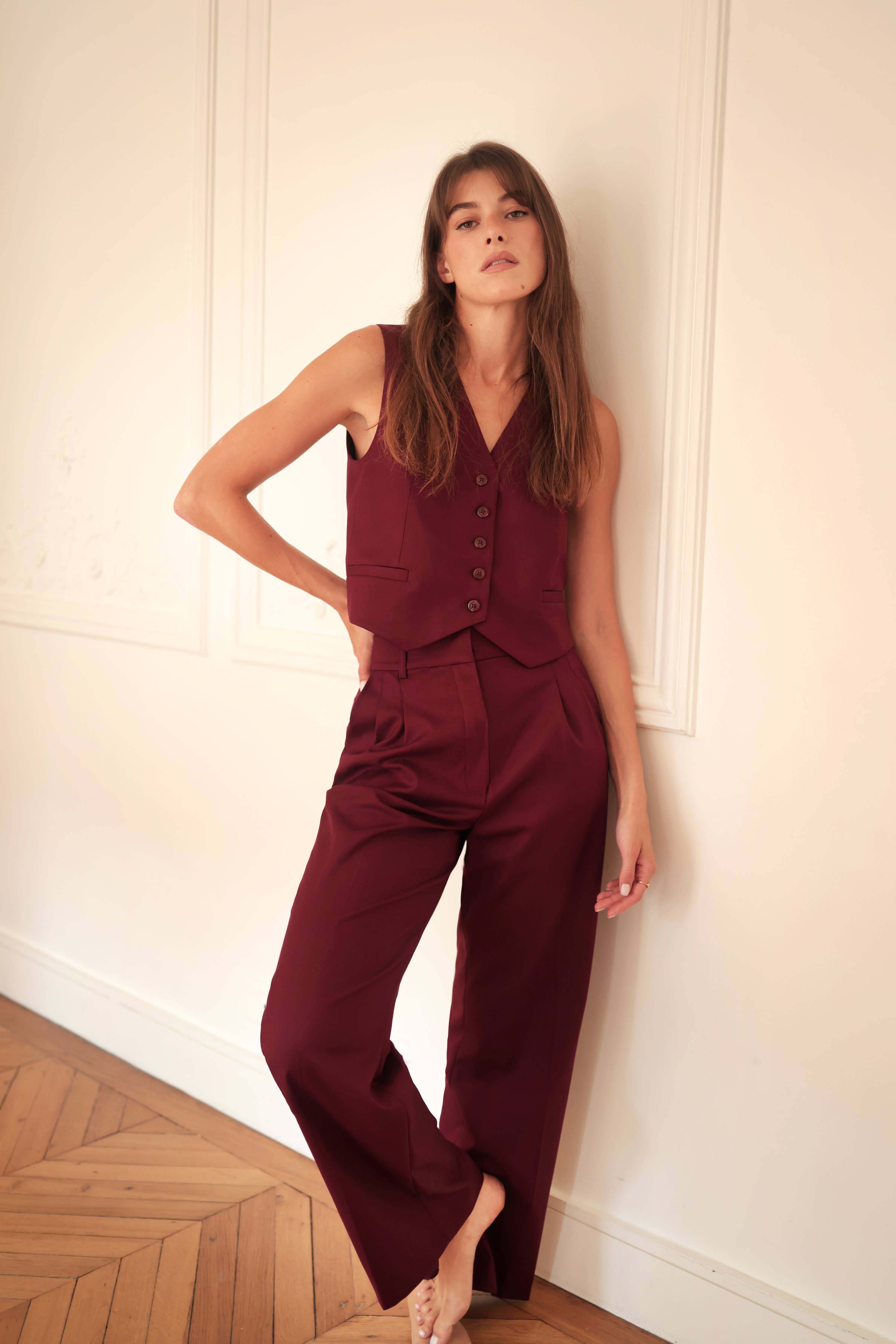 A woman standing against a wall, wearing burgundy sleeveless suit pants with a v-neckline and front pleats.