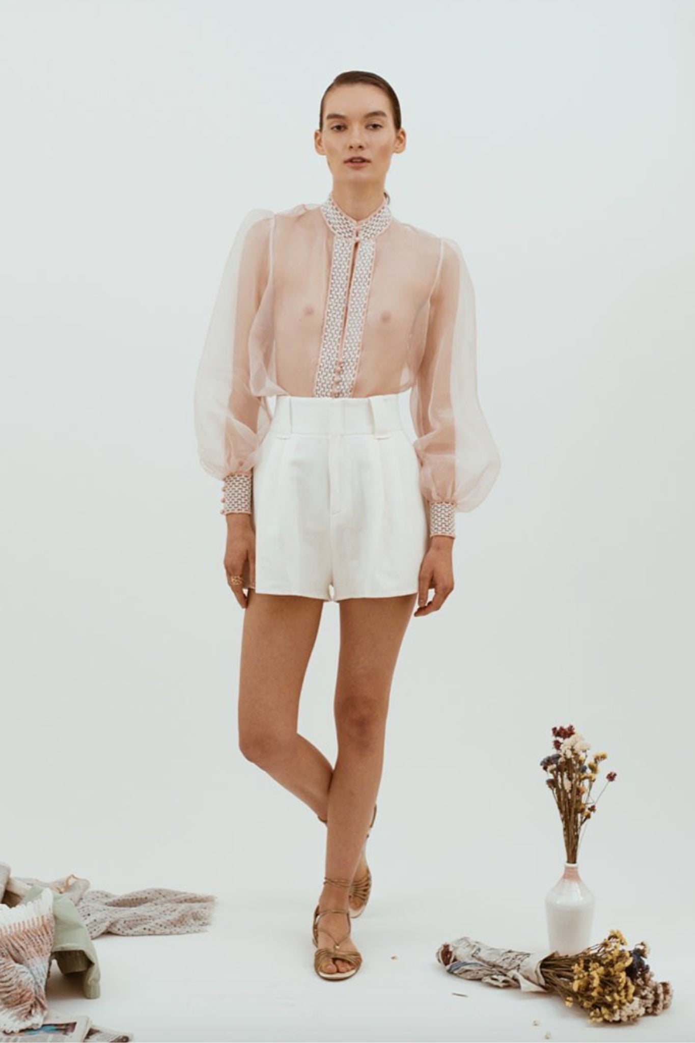 Woman wearing a sheer pink blouse with lace details and white shorts on a white background.