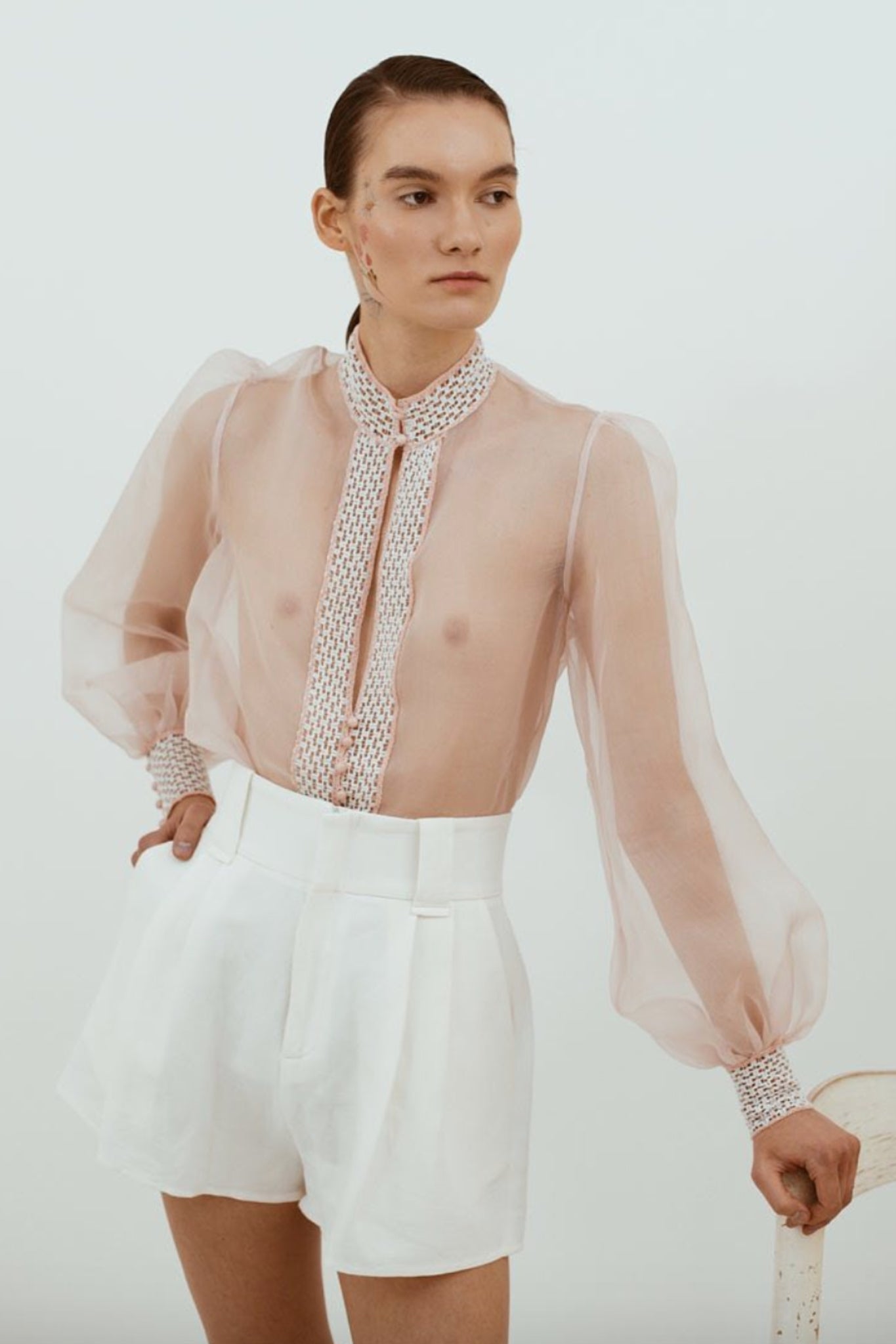 Model wearing a sheer top with a patterned scarf and white shorts on a plain background