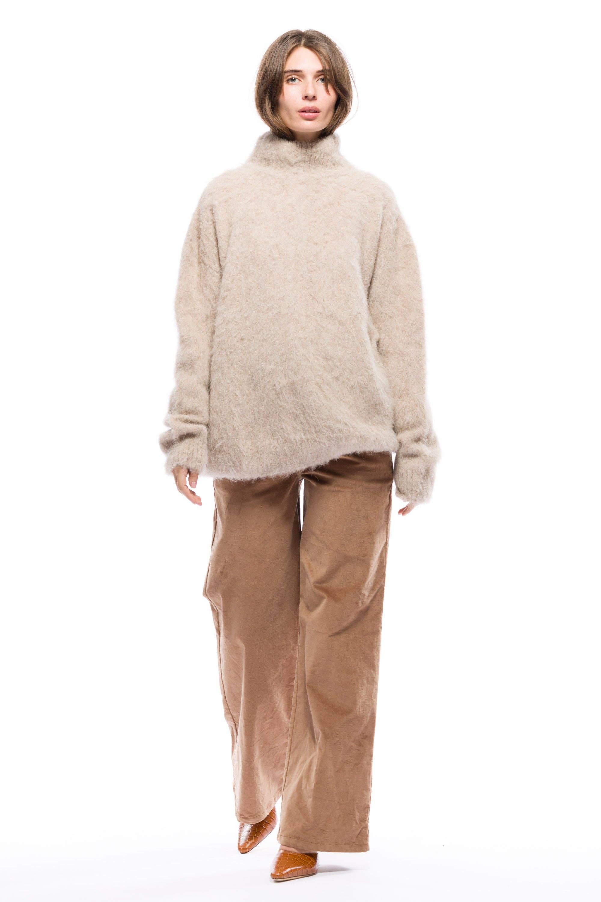 A person standing wearing a beige turtleneck sweater and brown pants.