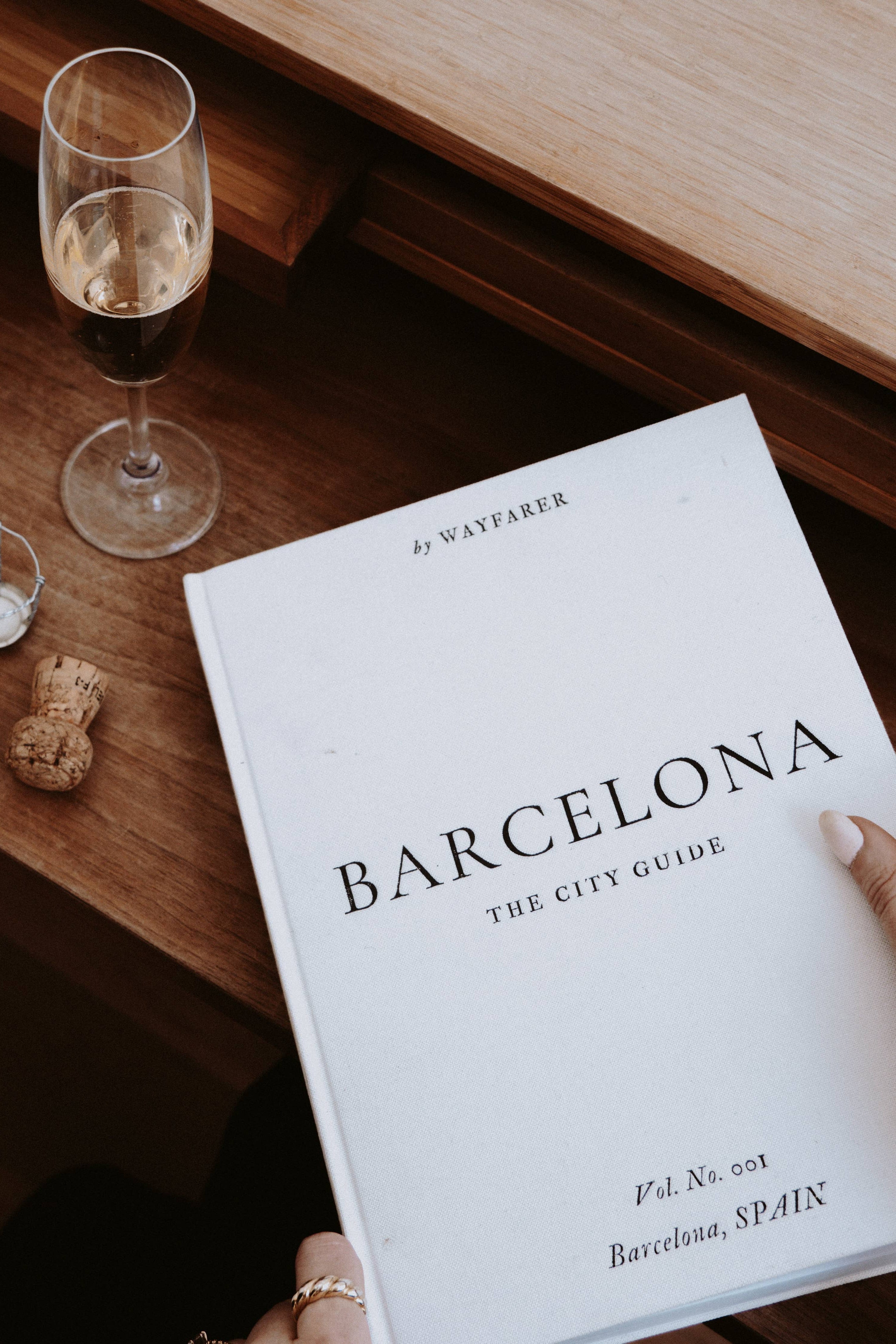 Book titled 'Barcelona: The City Guide' on a wooden table with a glass of wine.