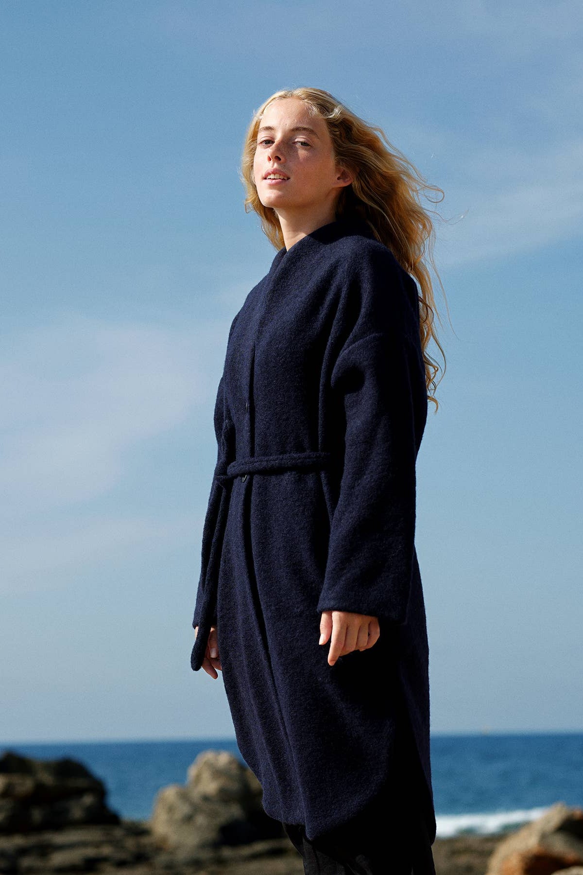 Woman wearing a navy blue coat standing on a rocky coastline with blue sky and ocean in the background