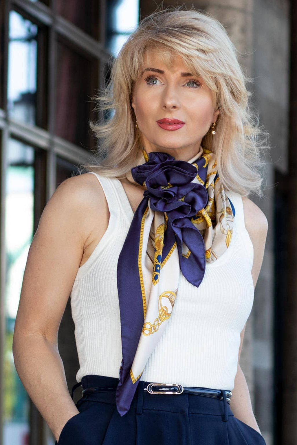 Woman wearing a white sleeveless top with a colorful scarf, standing against a stone wall.