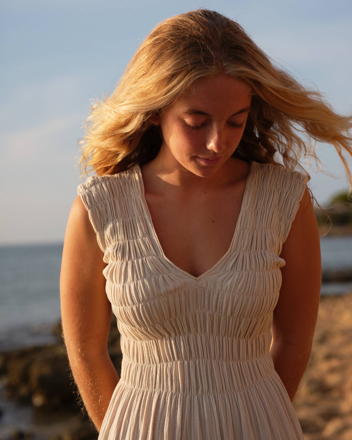 Woman in a beige dress standing on a beach with ocean and sky in the background