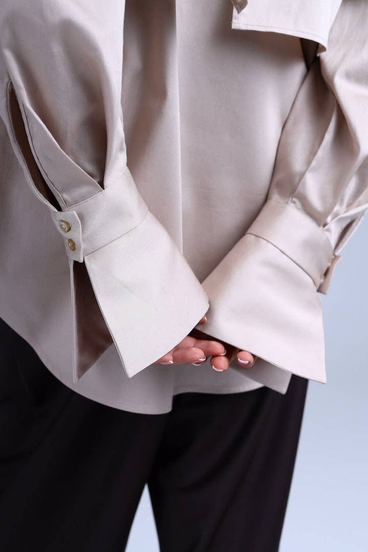 Close-up of a person wearing a beige blouse with rolled-up sleeves and black pants on a light blue background.