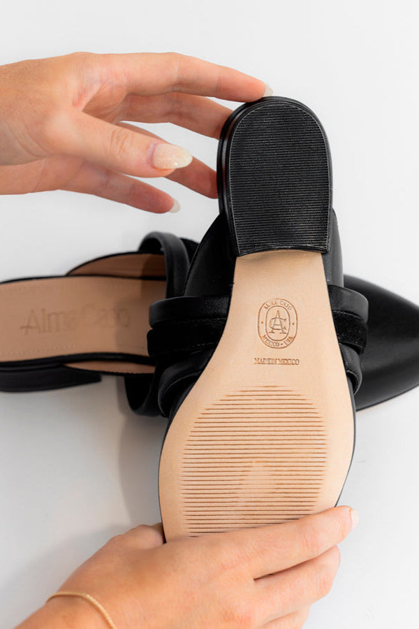 Black leather pointed-toe mule sandals shown from the bottom, with hands holding one sandal to display the tan rubber outsole and the embossed logo. A second sandal lies in the background on a clean white surface.