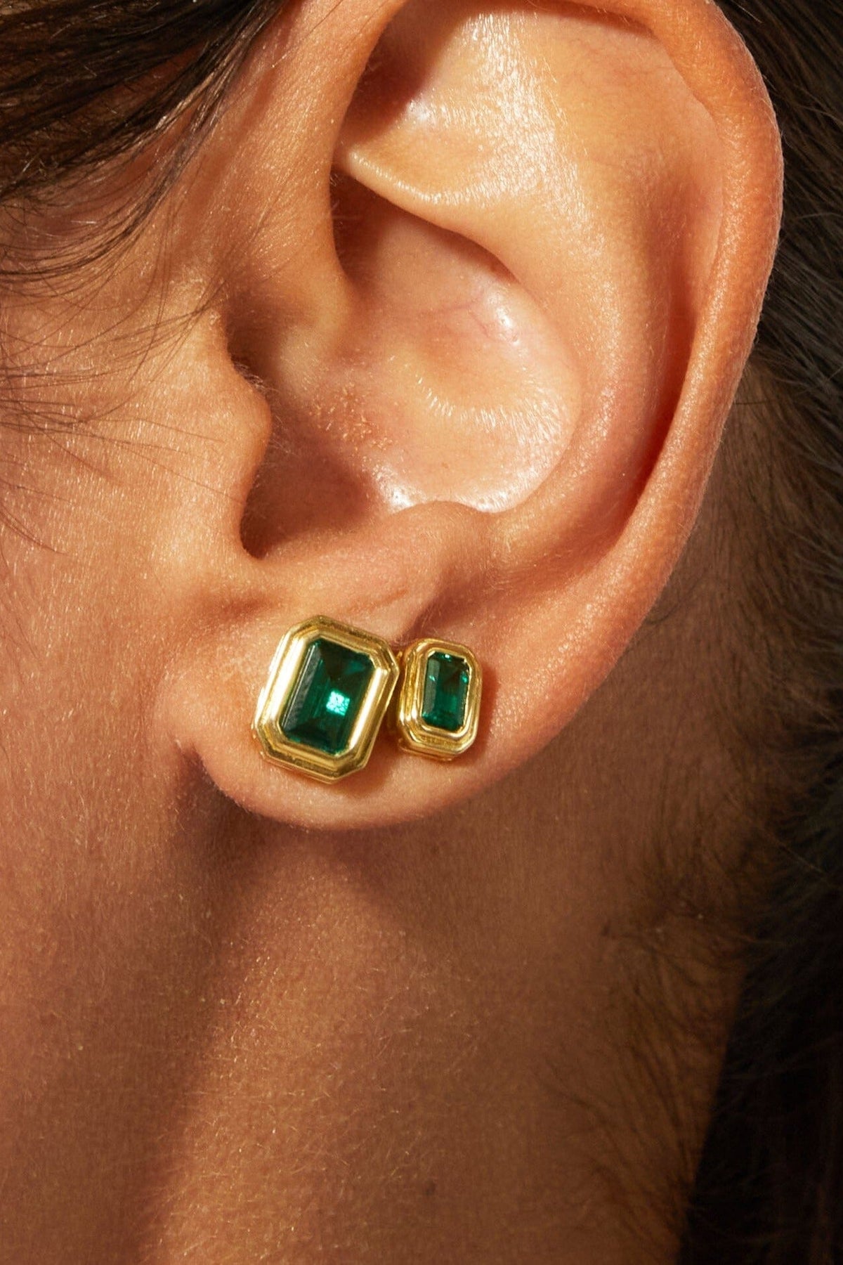 Close-up of an ear wearing gold and green emerald stud earrings.