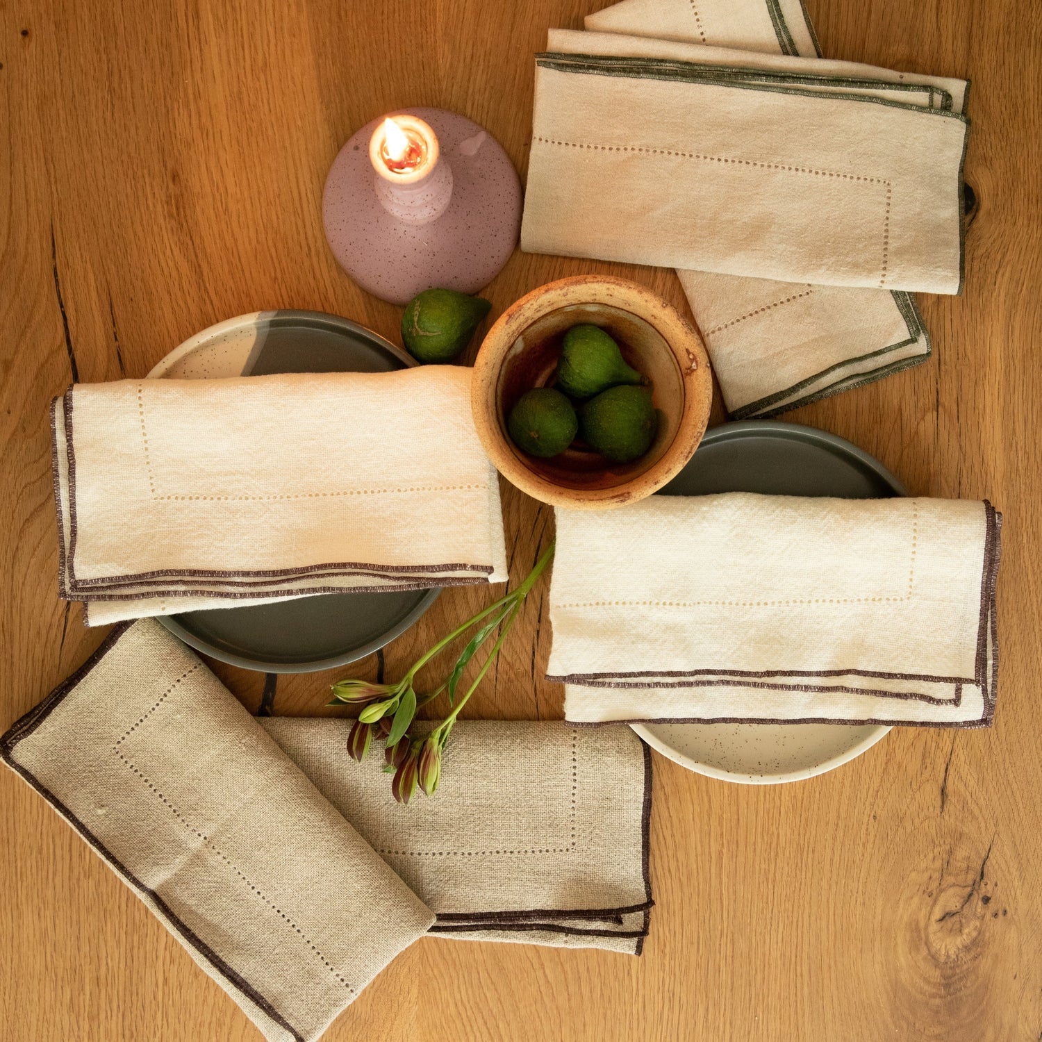 Beige napkins with brown trim on a wooden table with a candle and bowl of limes.