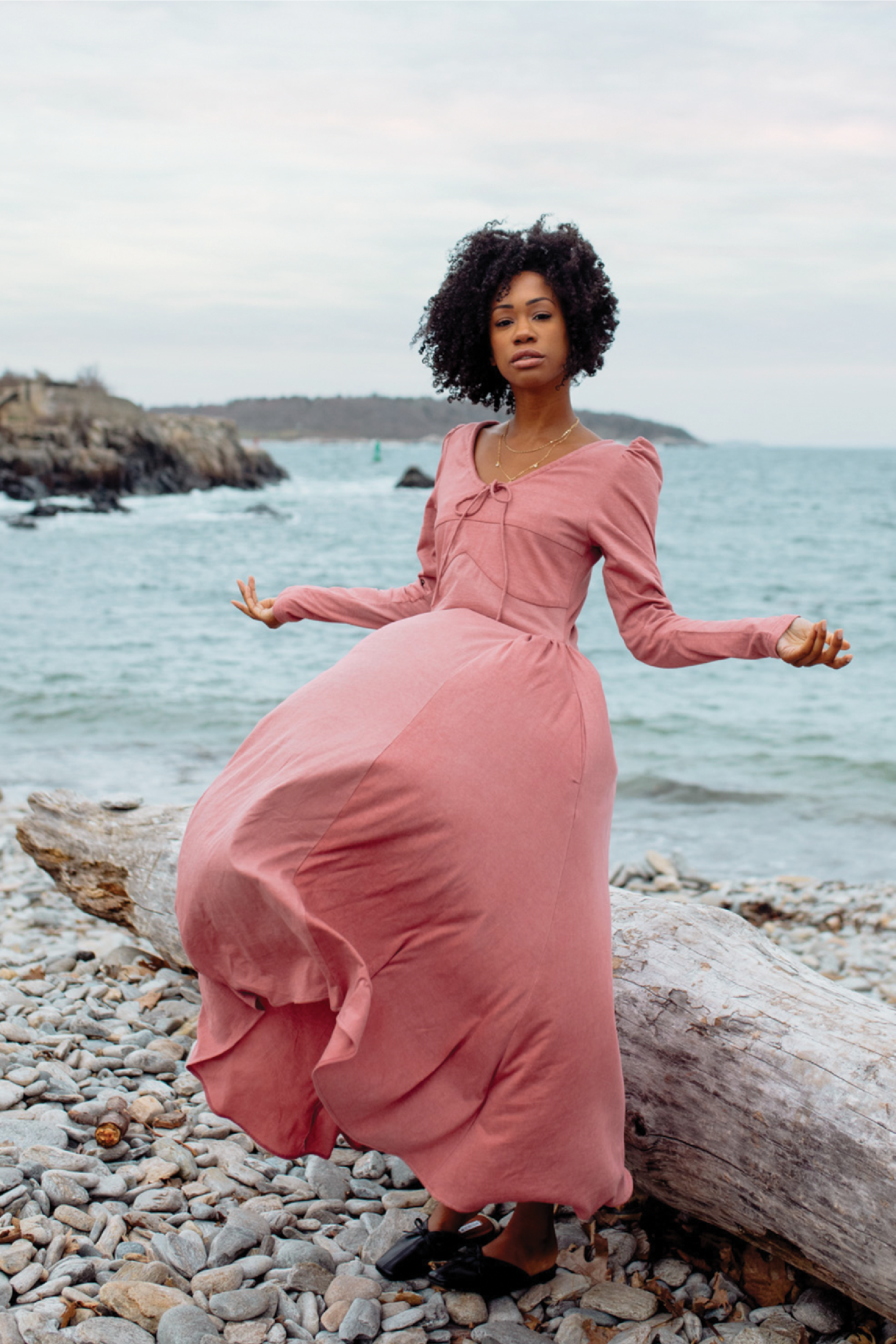 Woman in a pink dress standing on a rocky beach with ocean in the background