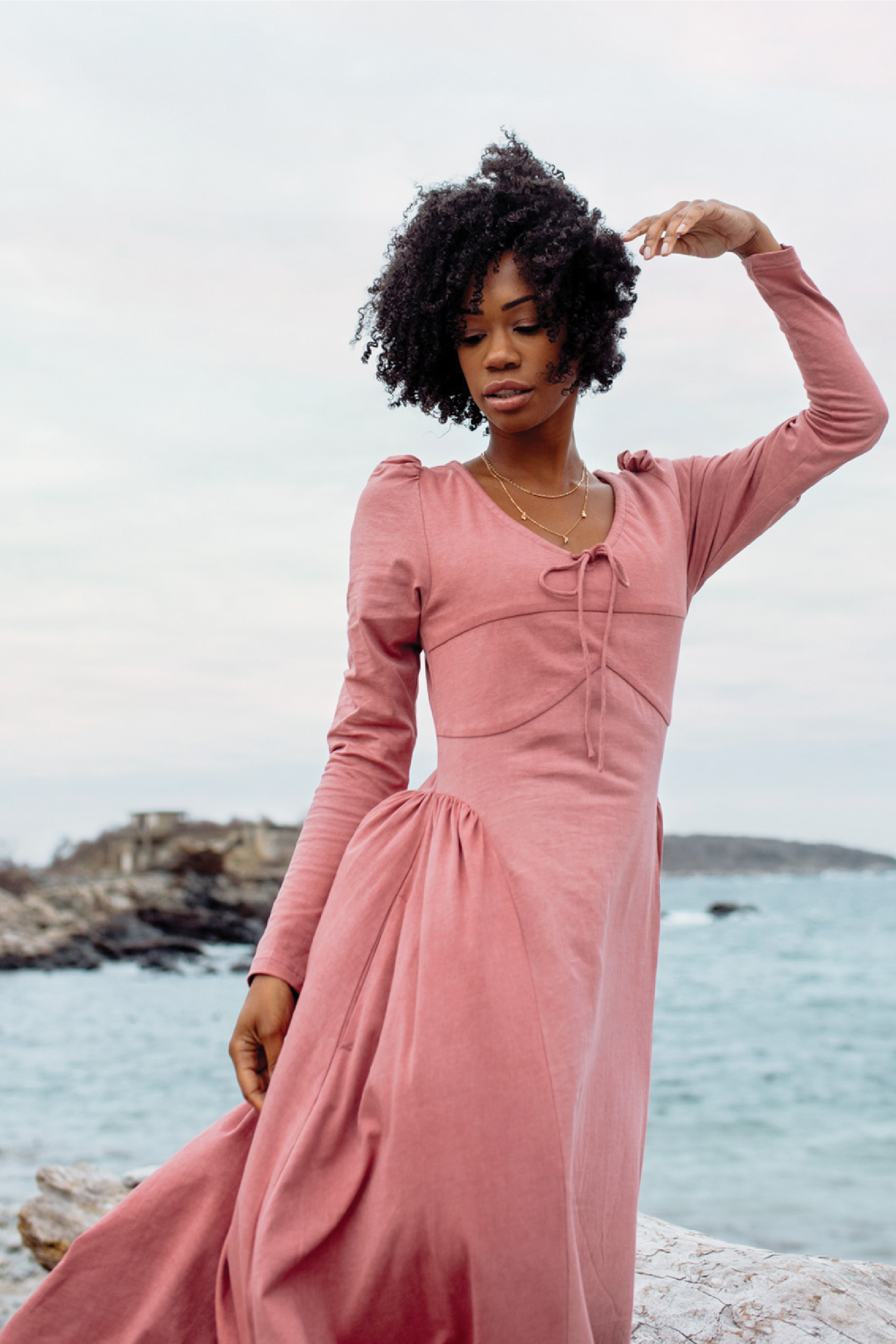 Woman in a pink dress standing on a rocky beach with ocean and sky in the background