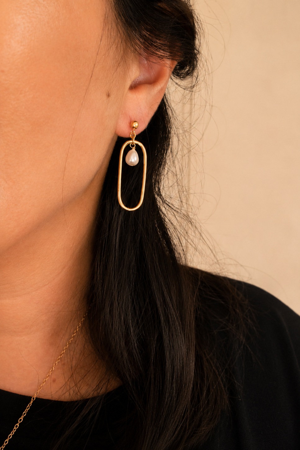 Close-up of a person wearing gold hoop earrings with a pearl accent against a beige background
