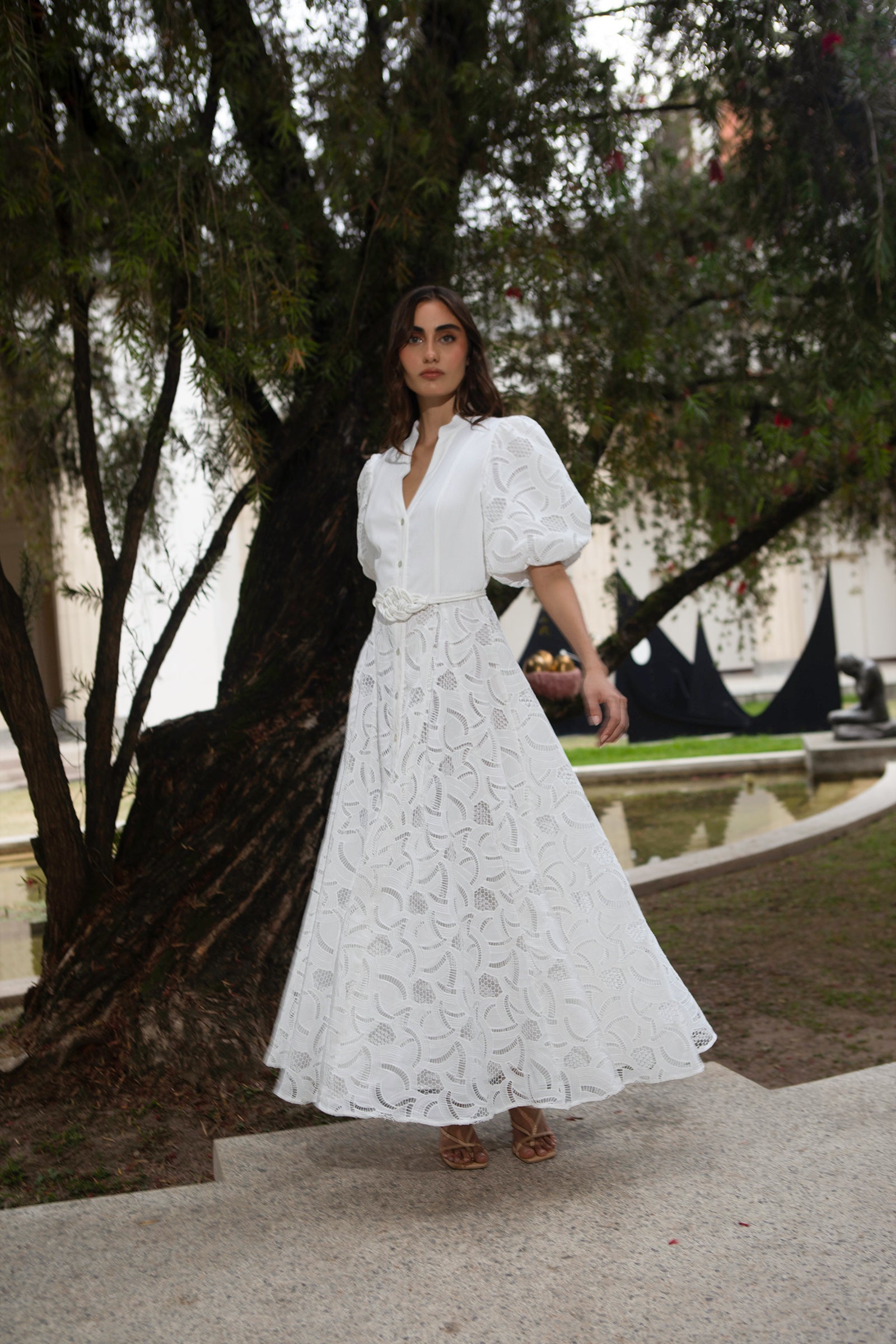 Woman in a white lace dress standing outdoors with trees and a fountain in the background