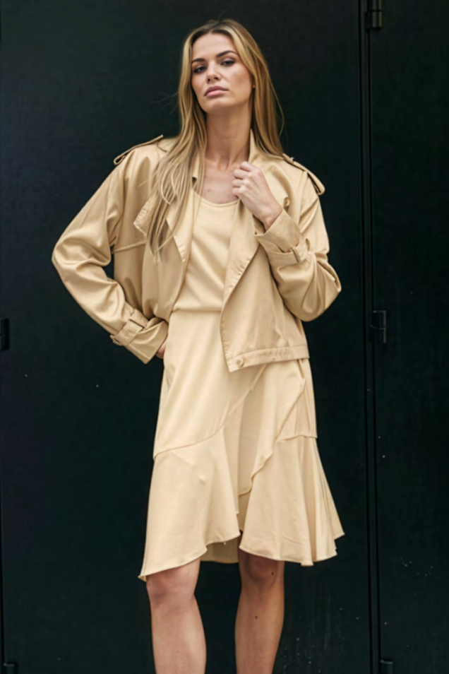 A woman modeling a beige satin-like jacket and matching dress against a dark black background.