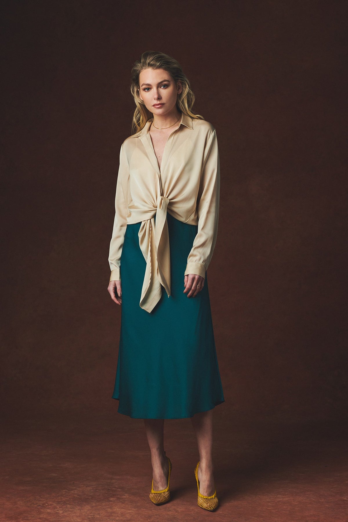 A woman stands against a dark brown studio backdrop wearing a cream satin-like long-sleeve shirt tied at the waist over a teal midi skirt, with yellow pointed heels.