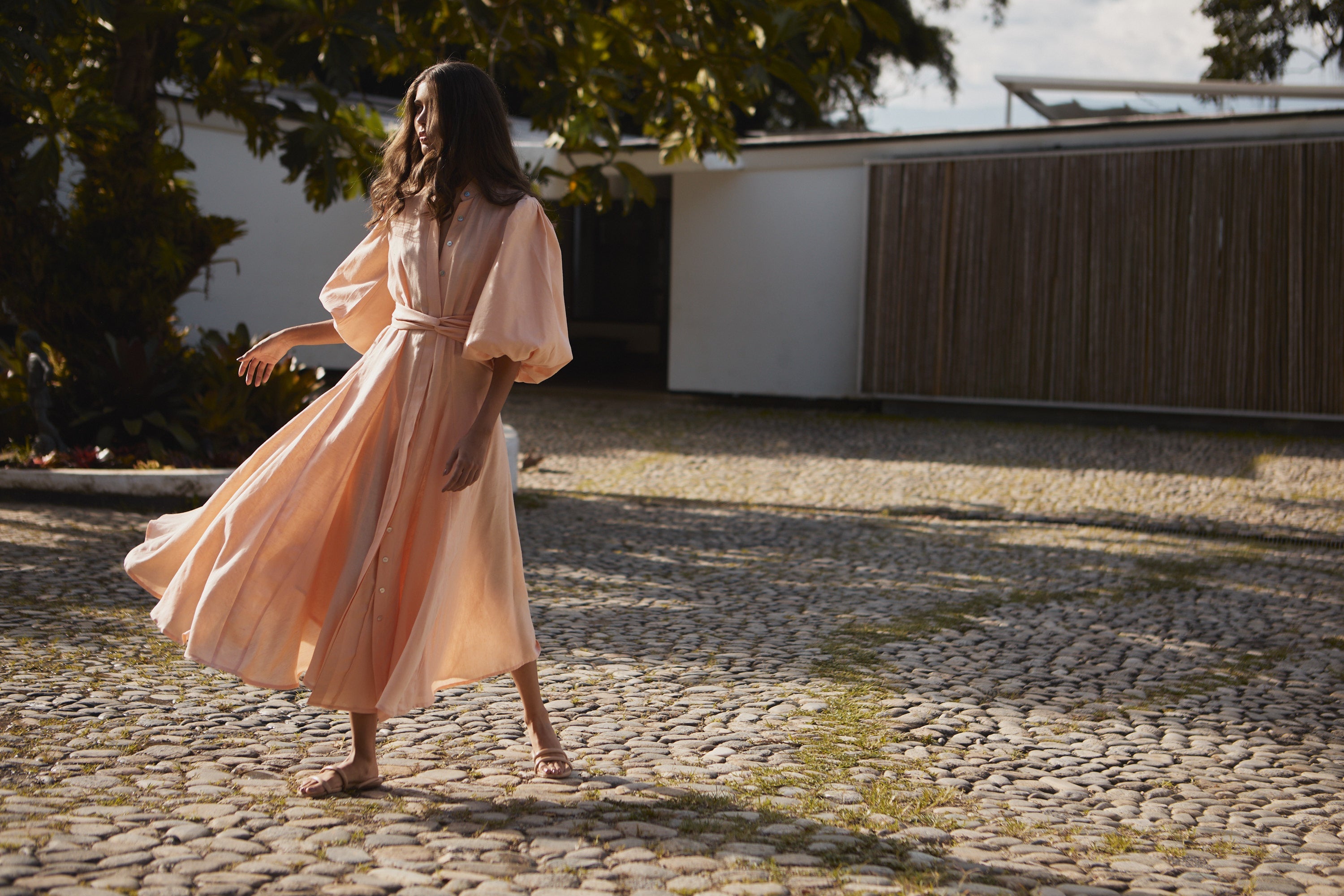 Woman in a flowing dress walking on a cobblestone path with trees and a building in the background.