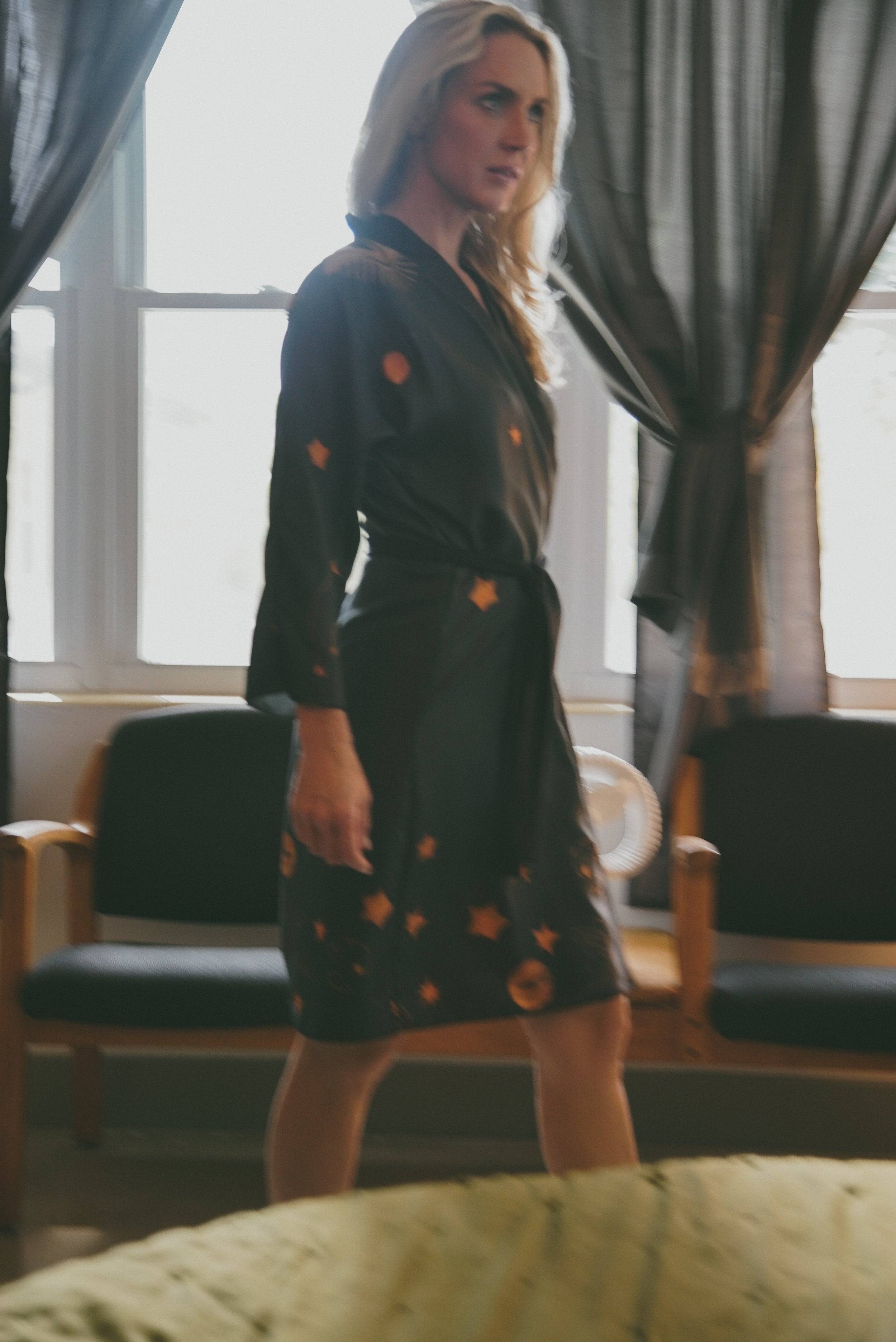 Woman in a dark dress with celestial patterns standing in a room with large windows and chairs.
