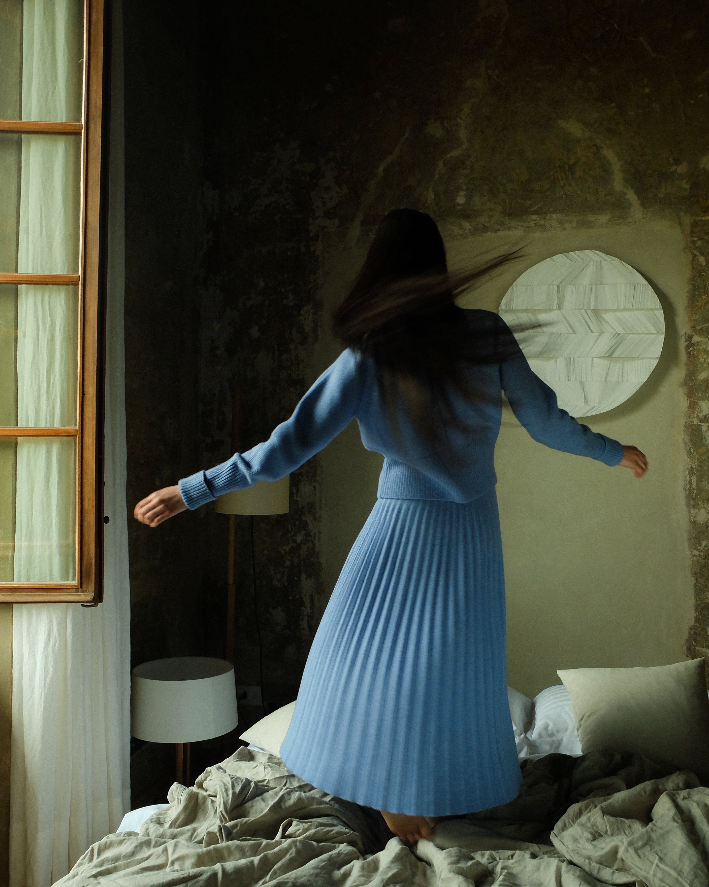 Woman in a blue dress standing in a bedroom with a textured wall and window.