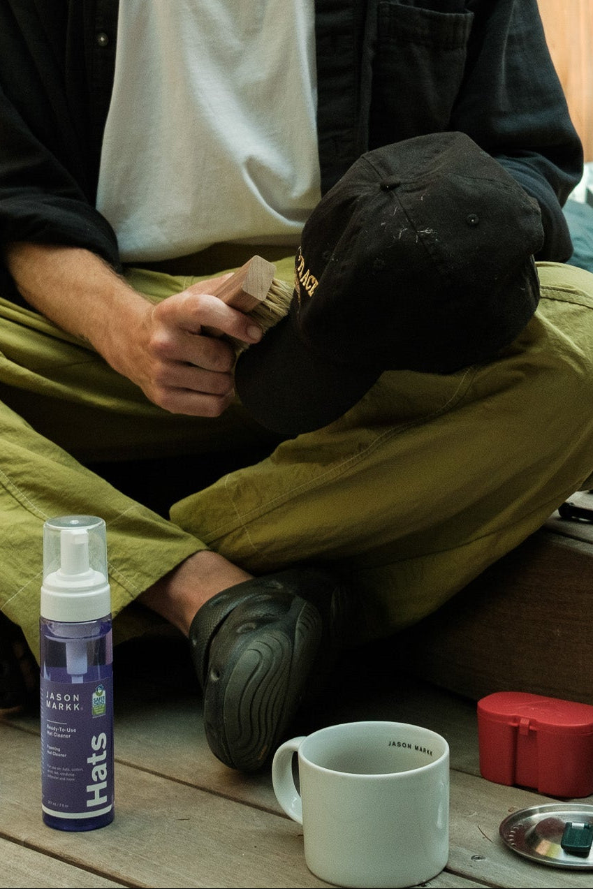 Man sitting on a wooden deck with camping gear and a cup, wearing a black jacket and green pants.