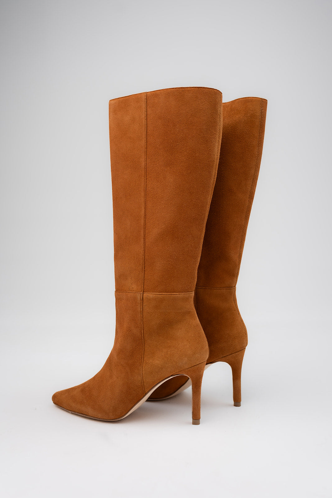 A pair of tall knee-high suede boots in a warm camel brown color, featuring a pointed toe and a slim high stiletto heel, photographed against a neutral light gray background.