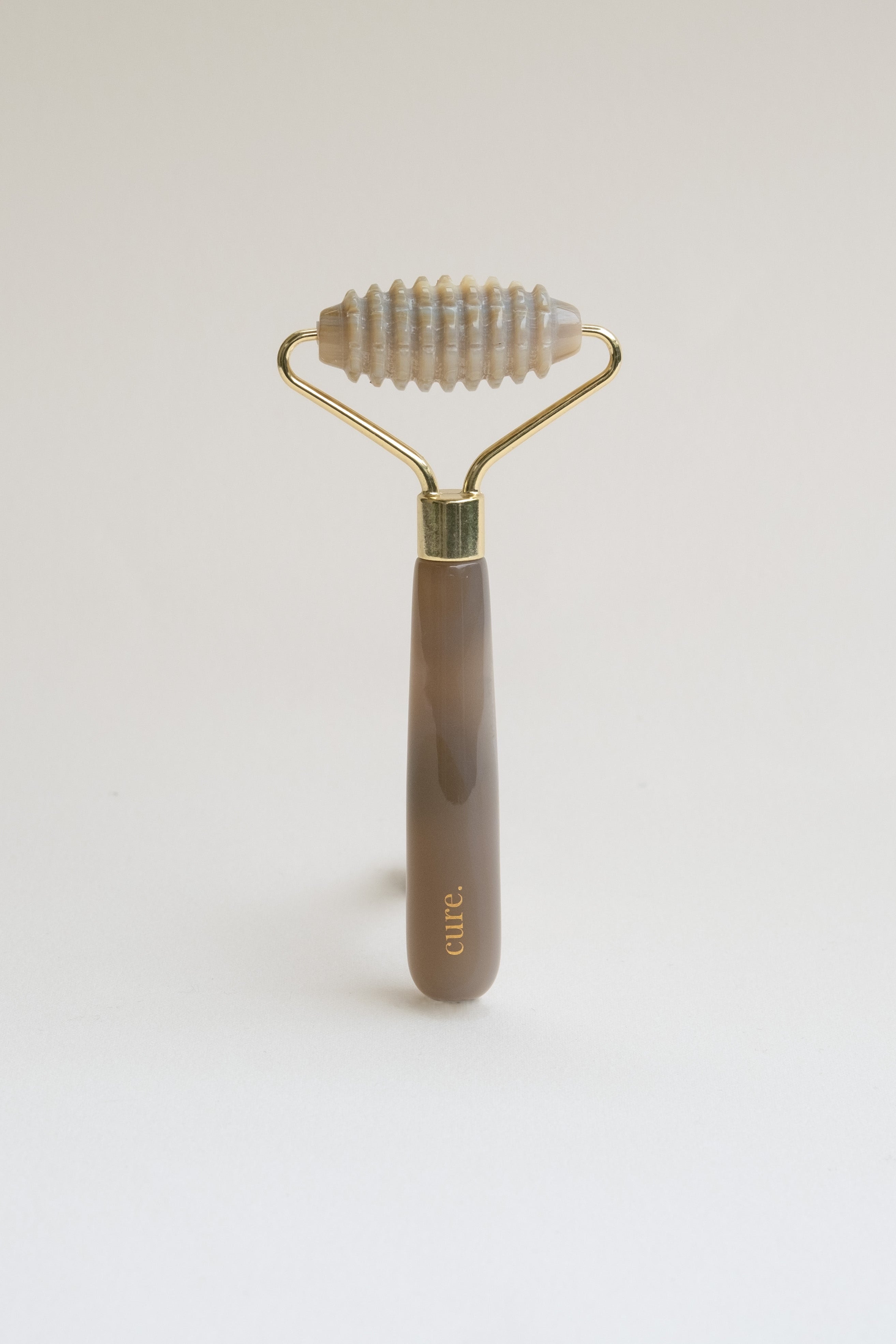 A facial roller tool with a marbled brown and gray handle, a gold metal frame, and a ribbed silicone roller head, standing upright on a light gray background. The brand name 'CELE' is printed in gold on the handle.