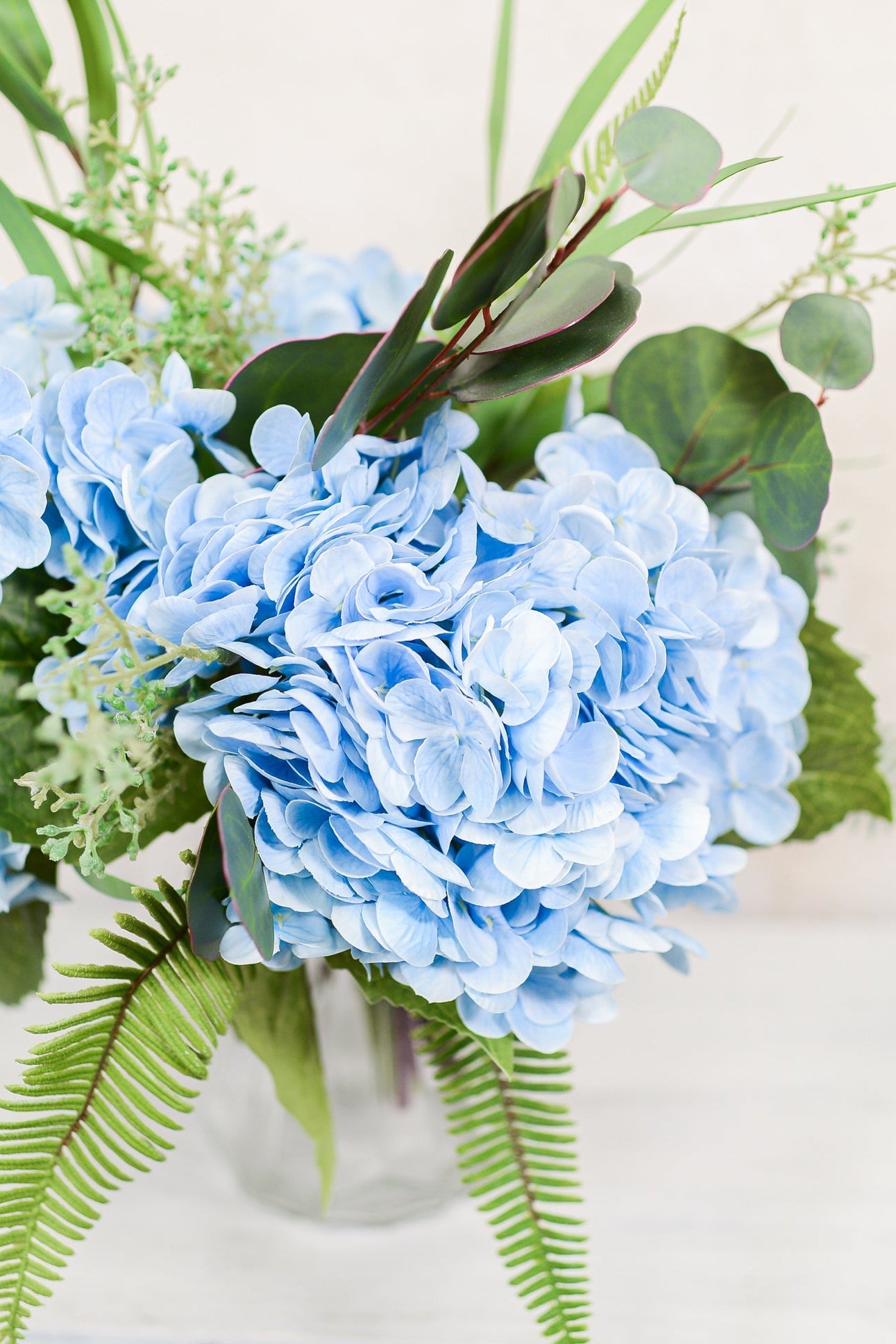 Bouquet of blue hydrangeas with greenery in a clear vase on a light background