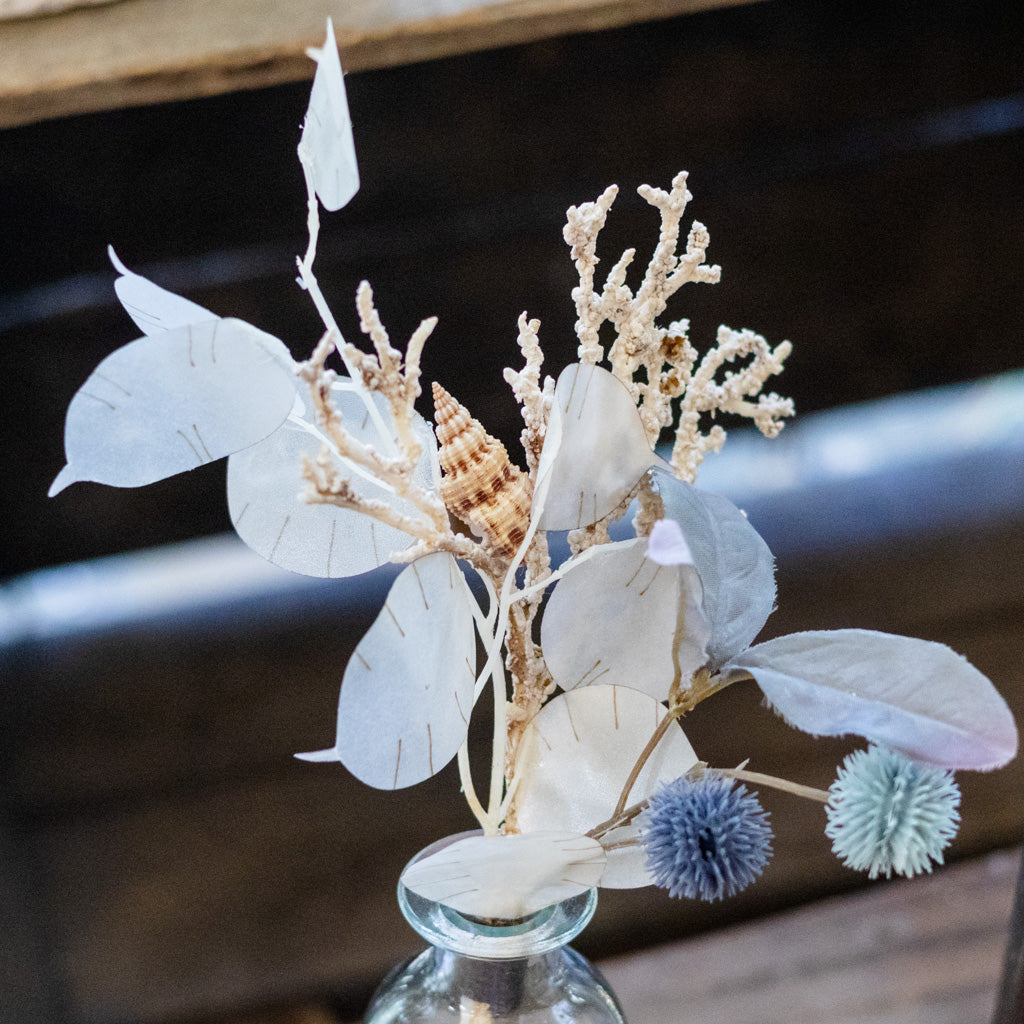 A dried floral arrangement featuring pale leaves, coral-like stems, a small seashell, and two pom-pom flowers in muted blue and green tones, displayed in a clear glass vase against a dark wooden background.