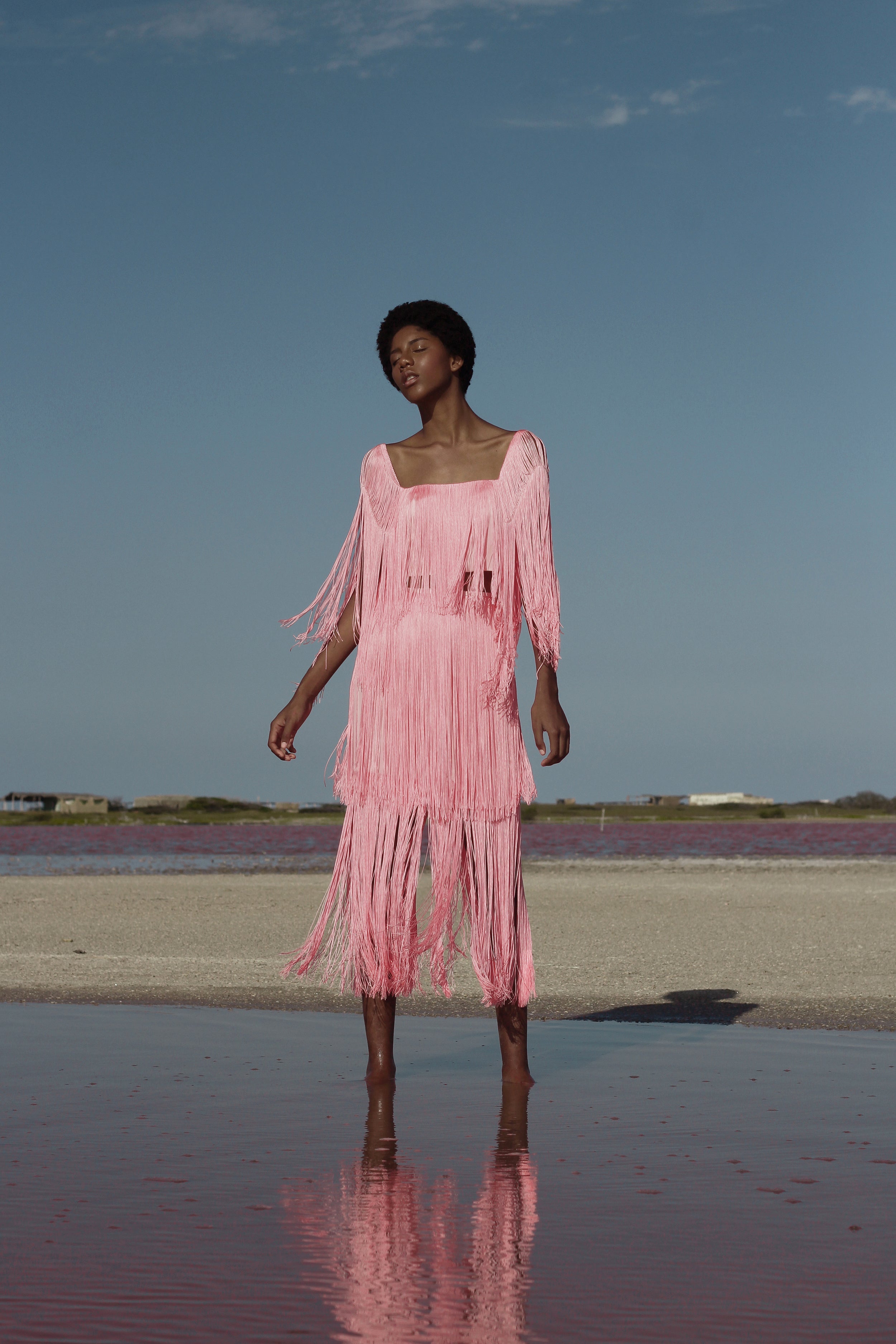 Woman in a pink dress standing on a reflective surface with a clear blue sky.