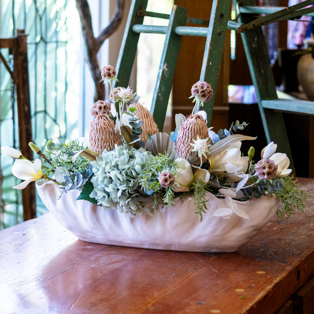 Decorative floral arrangement in a white shell on a wooden table with a green ladder in the background.