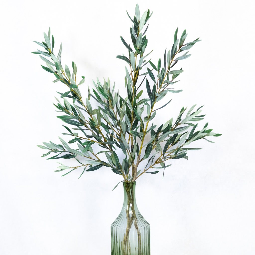 Artificial olive branch stem arrangement in a ribbed clear glass vase against a plain white background.