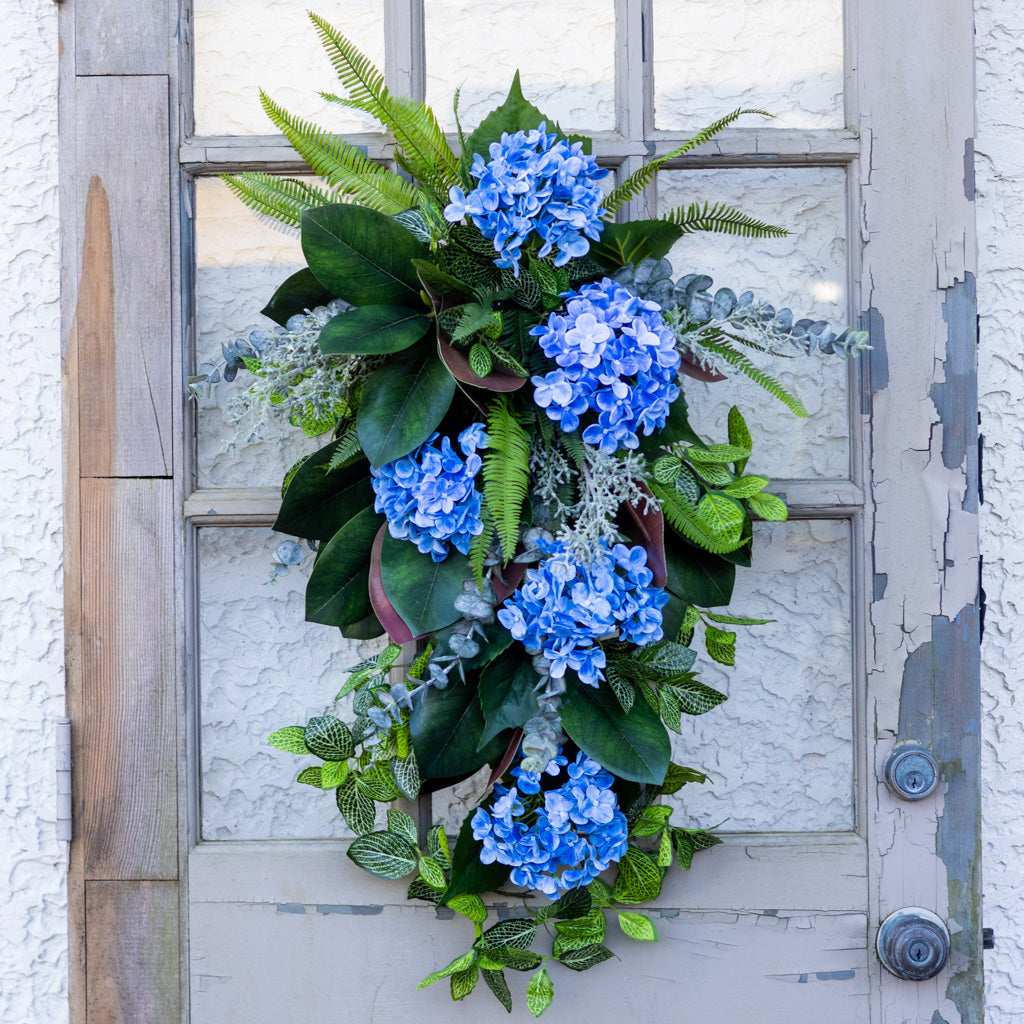 A decorative hanging floral arrangement with clusters of bright blue hydrangeas, green ferns, and leafy vines, displayed on a distressed gray wooden door.