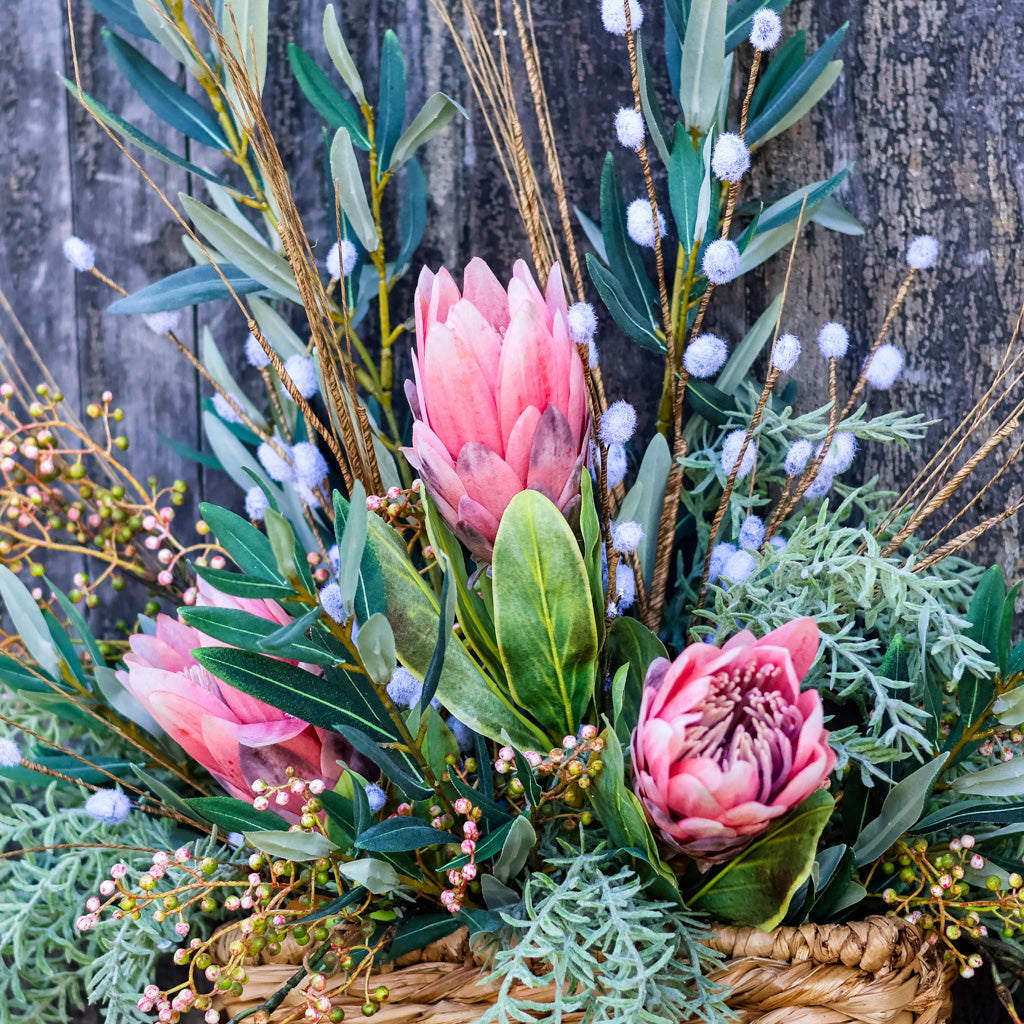 A rustic floral arrangement in a woven basket featuring pink protea-like flowers, green leaves, soft silvery foliage, and white fluffy buds, set against a weathered wood background.