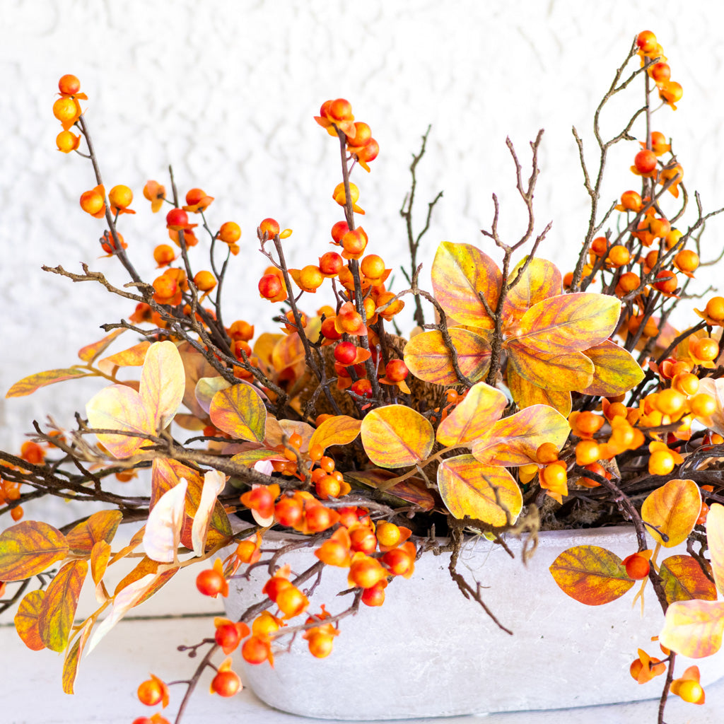 Autumn berry garland with orange and yellow leaves on brown branches, arranged in a white textured pot against a white wall.