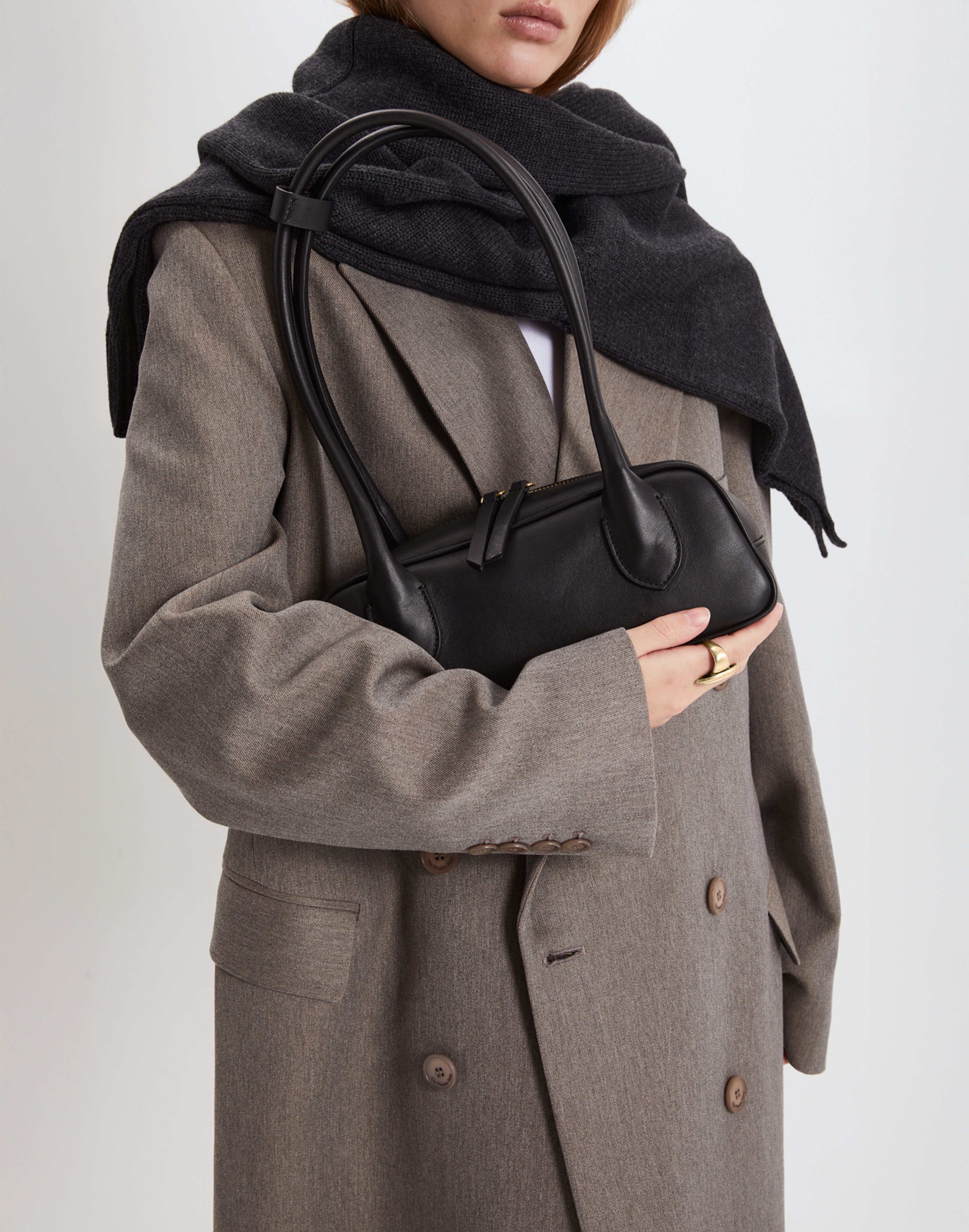 Person wearing a gray coat and black scarf holding a black handbag against a light background