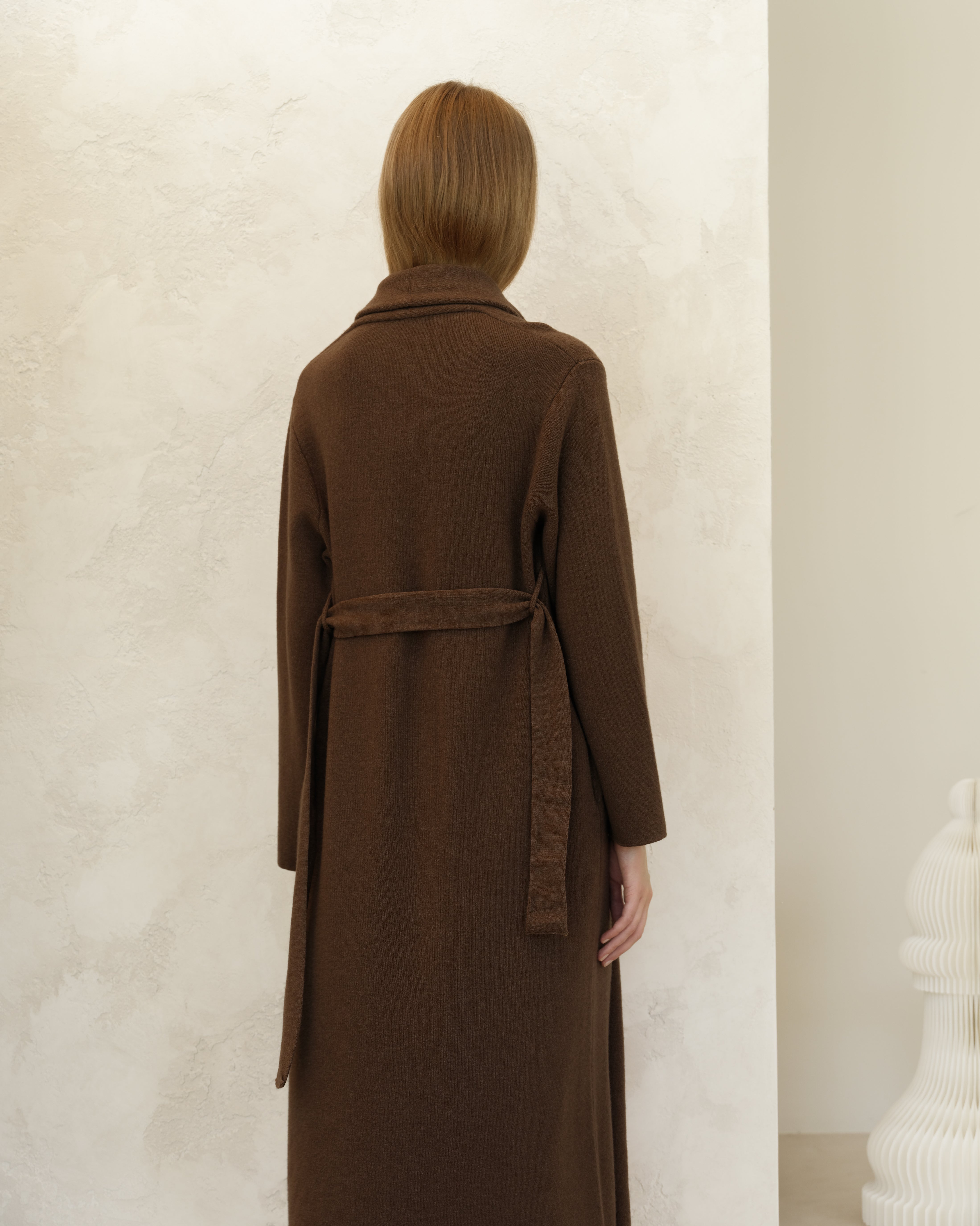 Person wearing a brown coat standing against a plain background