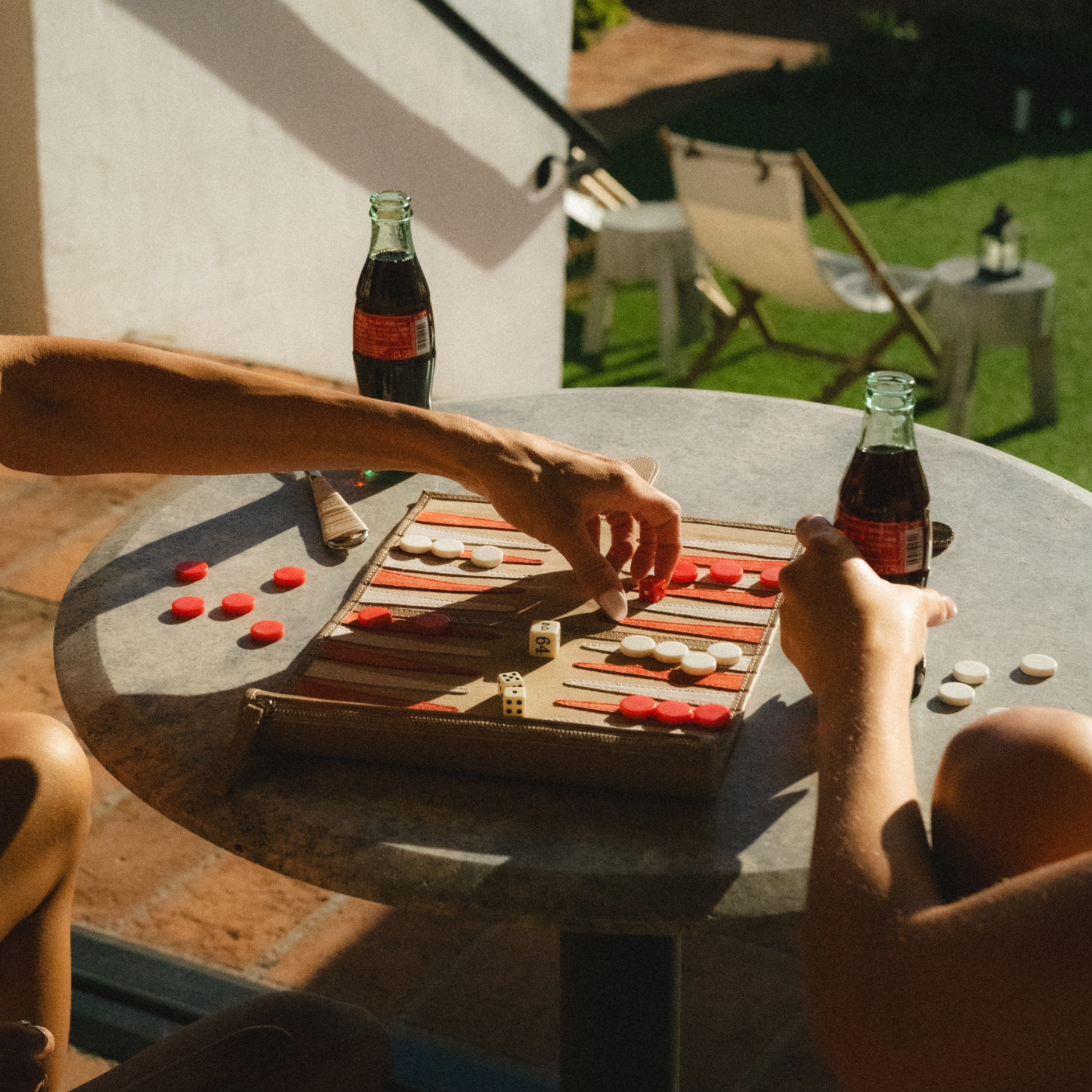 Two people playing backgammon at a table outdoors with Coca-Cola bottles.