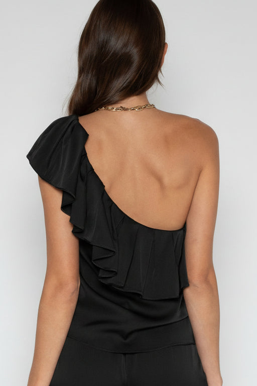 A woman shown from the back wearing a black one-shoulder ruffled top or dress against a plain light gray studio background.