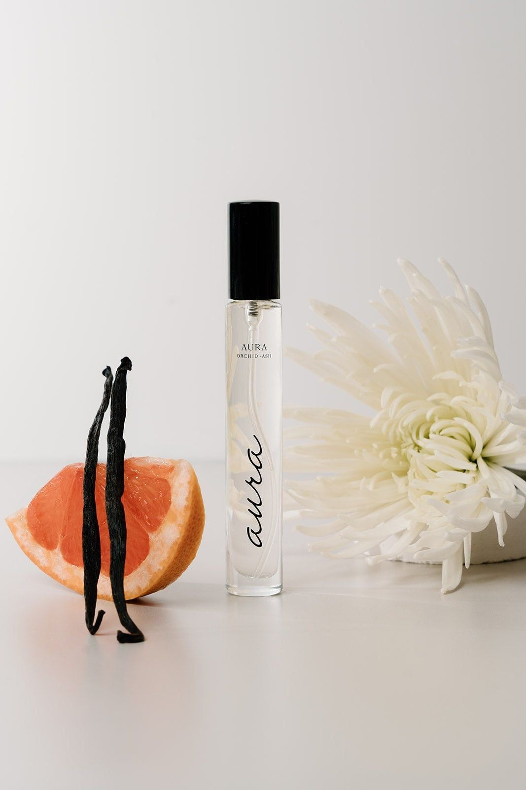 Perfume bottle with vanilla beans, grapefruit, and a white flower on a light background