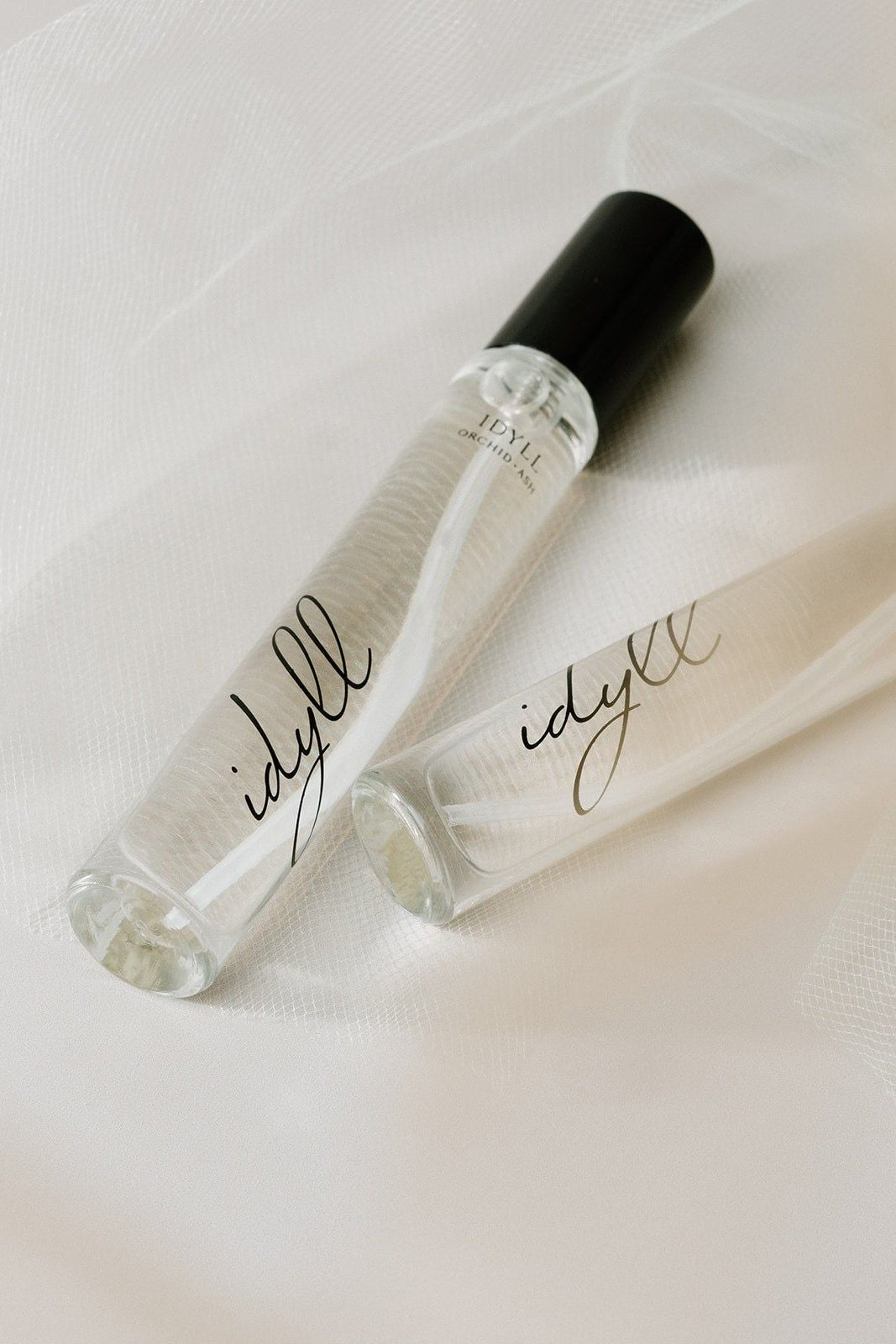 Two clear 'idyll' perfume bottles with black caps on a light fabric background