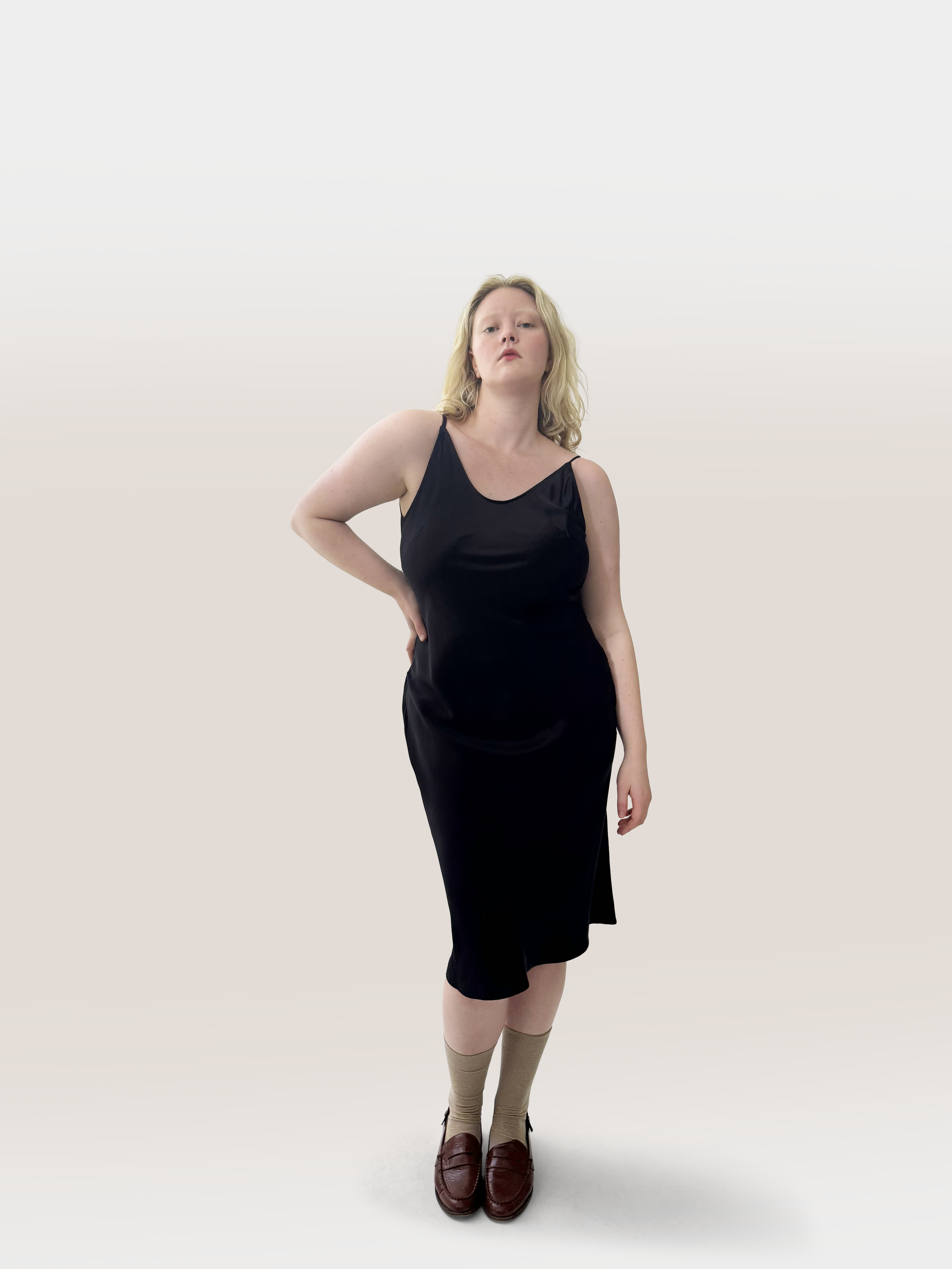 A woman modeling a black silk sleeveless midi dress with a scoop neck, paired with beige socks and dark brown loafers, standing against a light gray studio background.