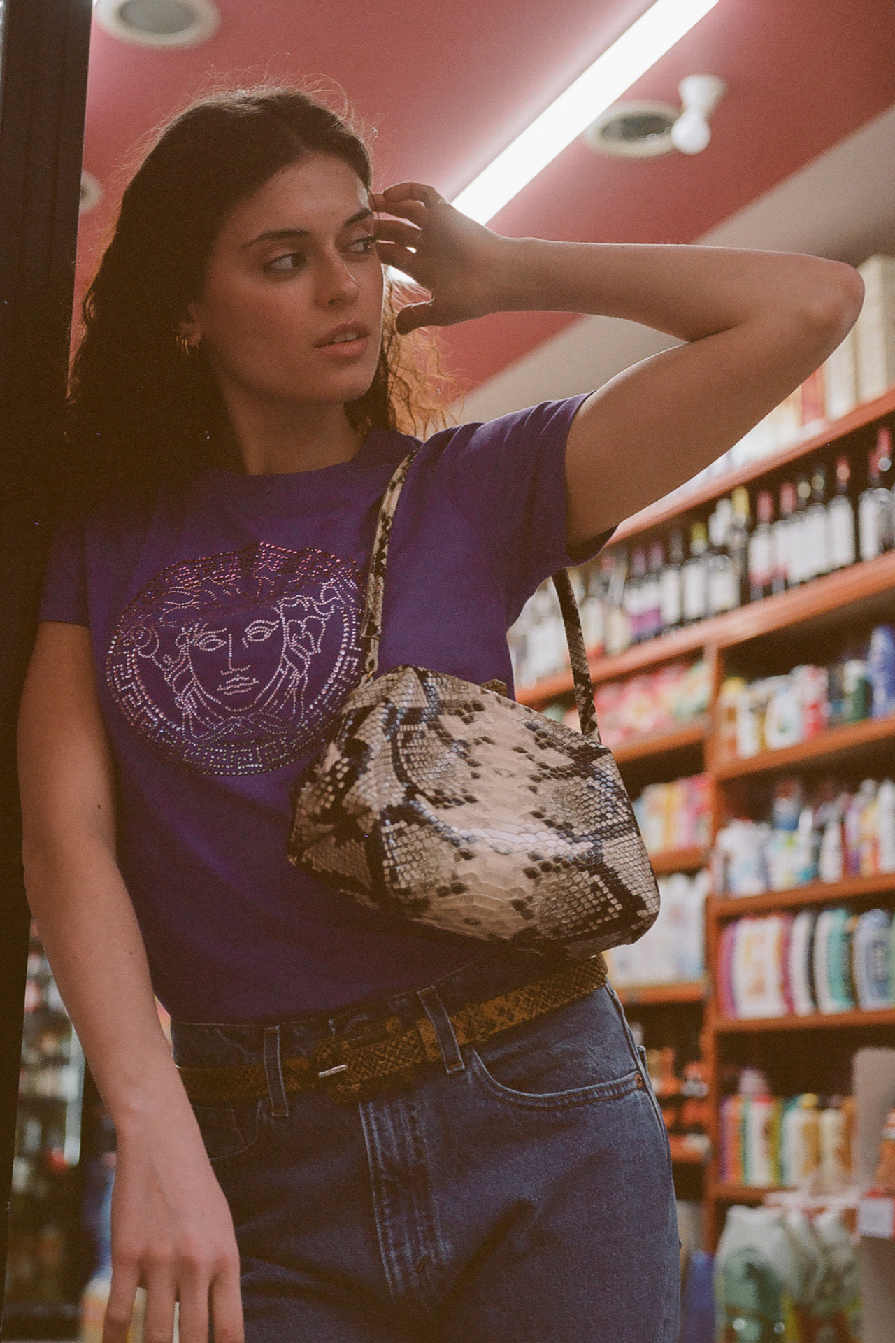 Woman in a store with shelves stocked with products, wearing a purple t-shirt, blue jeans, and a python print leather shoulder bag.