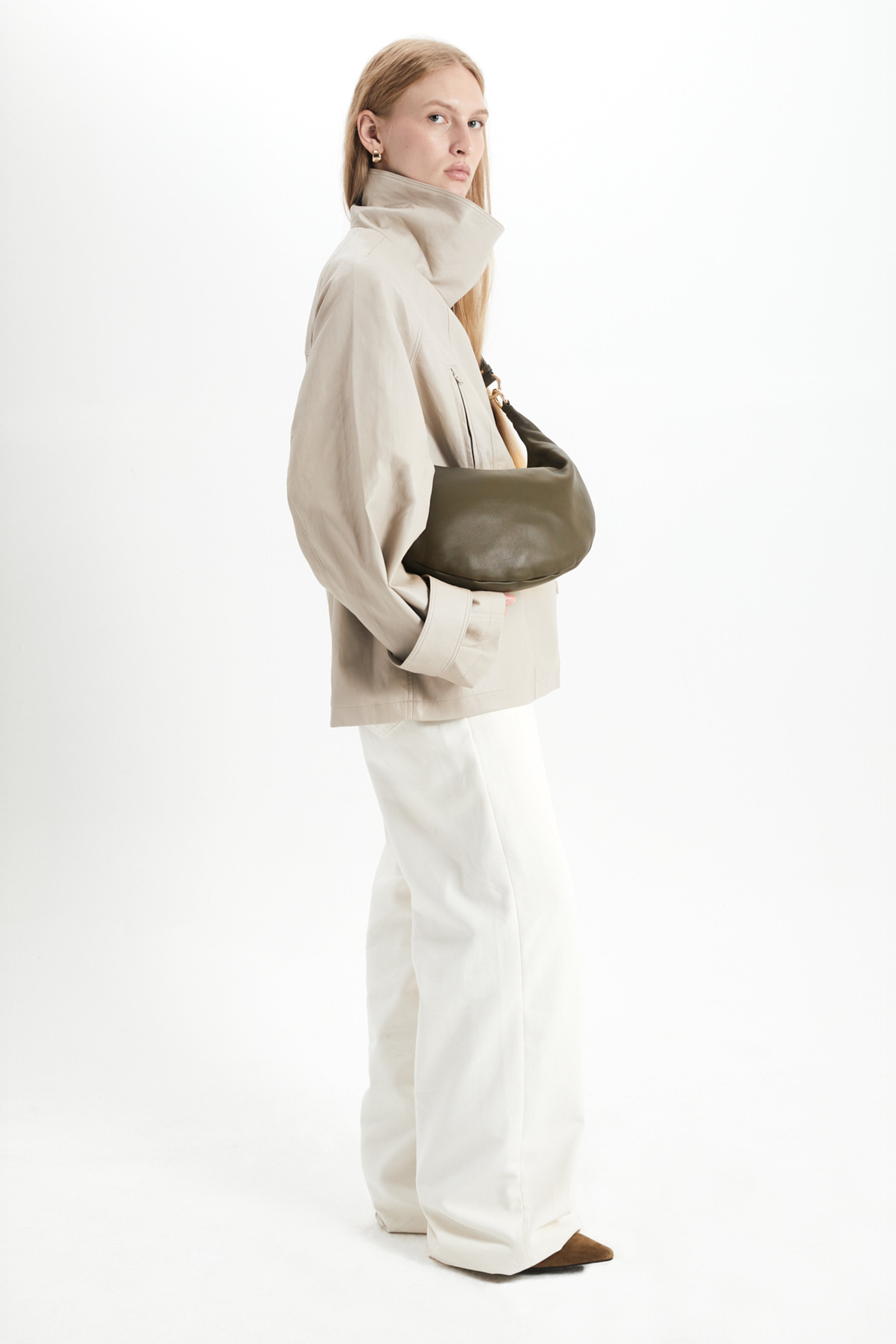 Woman holding a green handbag wearing a beige jacket and white pants on a light gray background