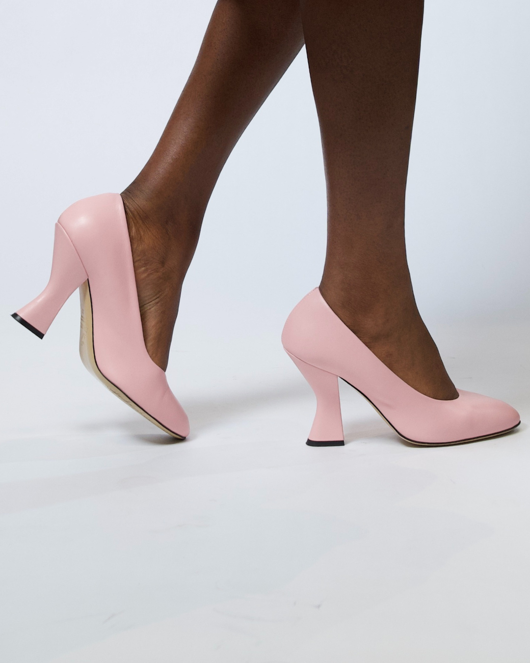 A pair of light pink pointed-toe high heels with kitten heels, shown on a model's feet against a light gray studio background.